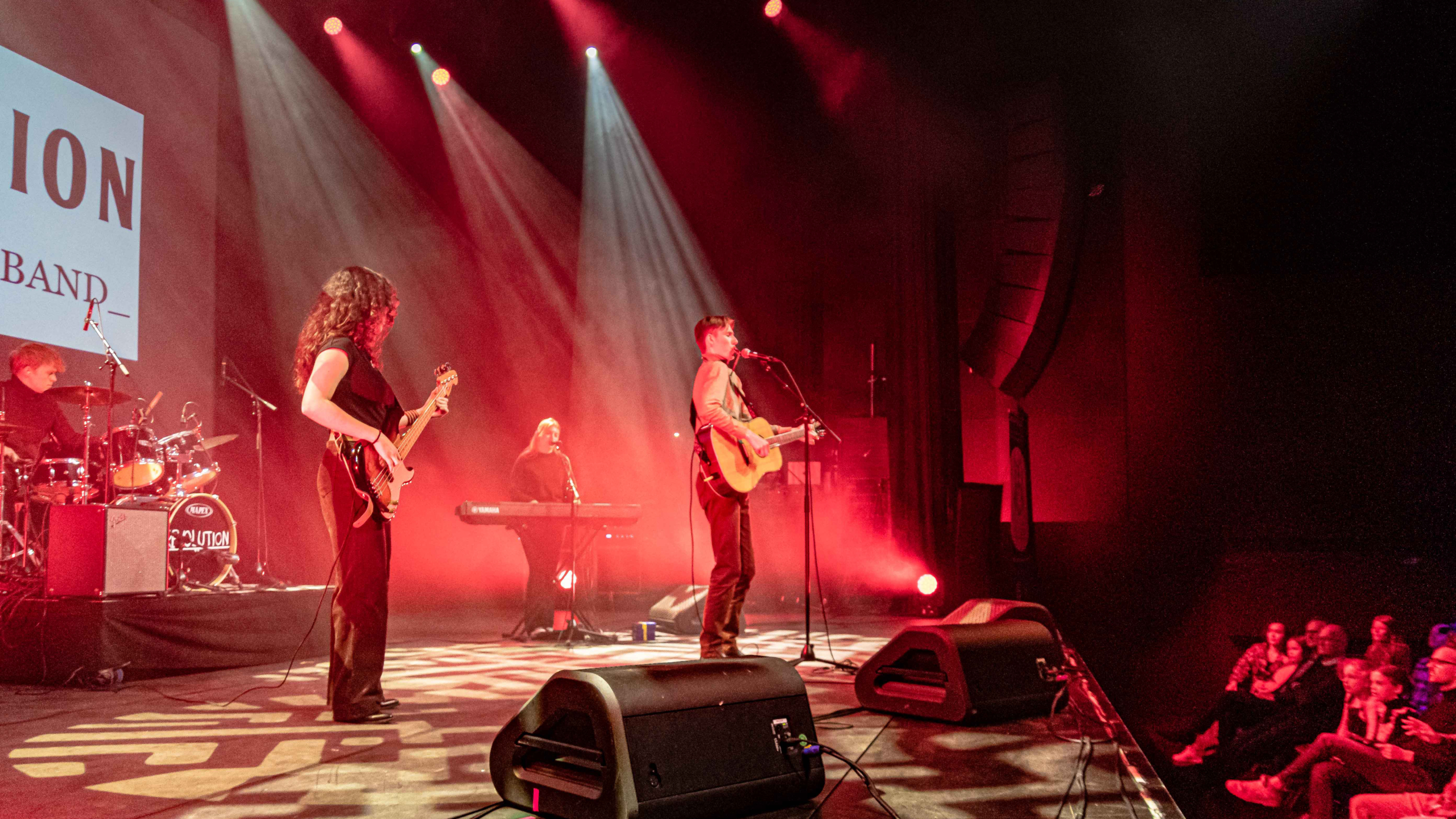 Muzikanten optreden op een podium verlicht met rood licht, inclusief een drummer, bassist, toetsenist en gitarist-zanger, met publiek dat toekijkt vanuit de stoelen.