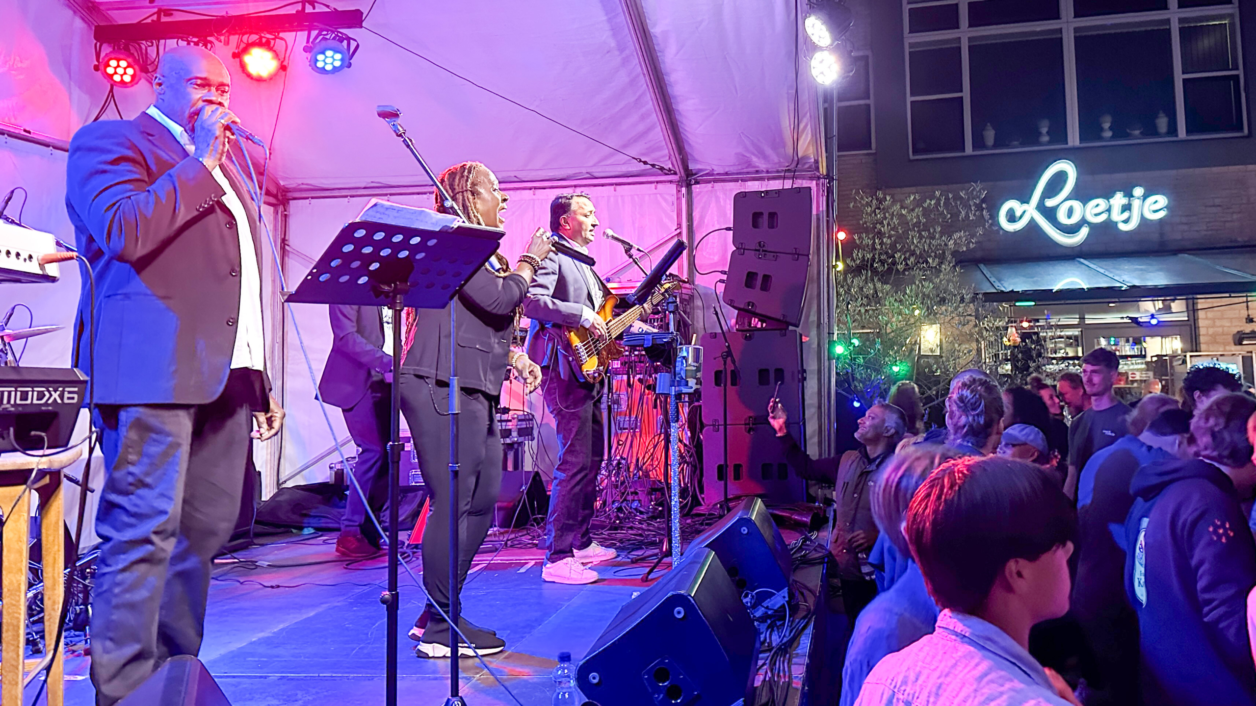 Een liveband optredend op een podium in een tent, met een zanger, zangeres en bassist, omringd door kleurrijke verlichting en een publiek vooraan. Op de achtergrond is een gebouw met de naam 'Loetje' zichtbaar.