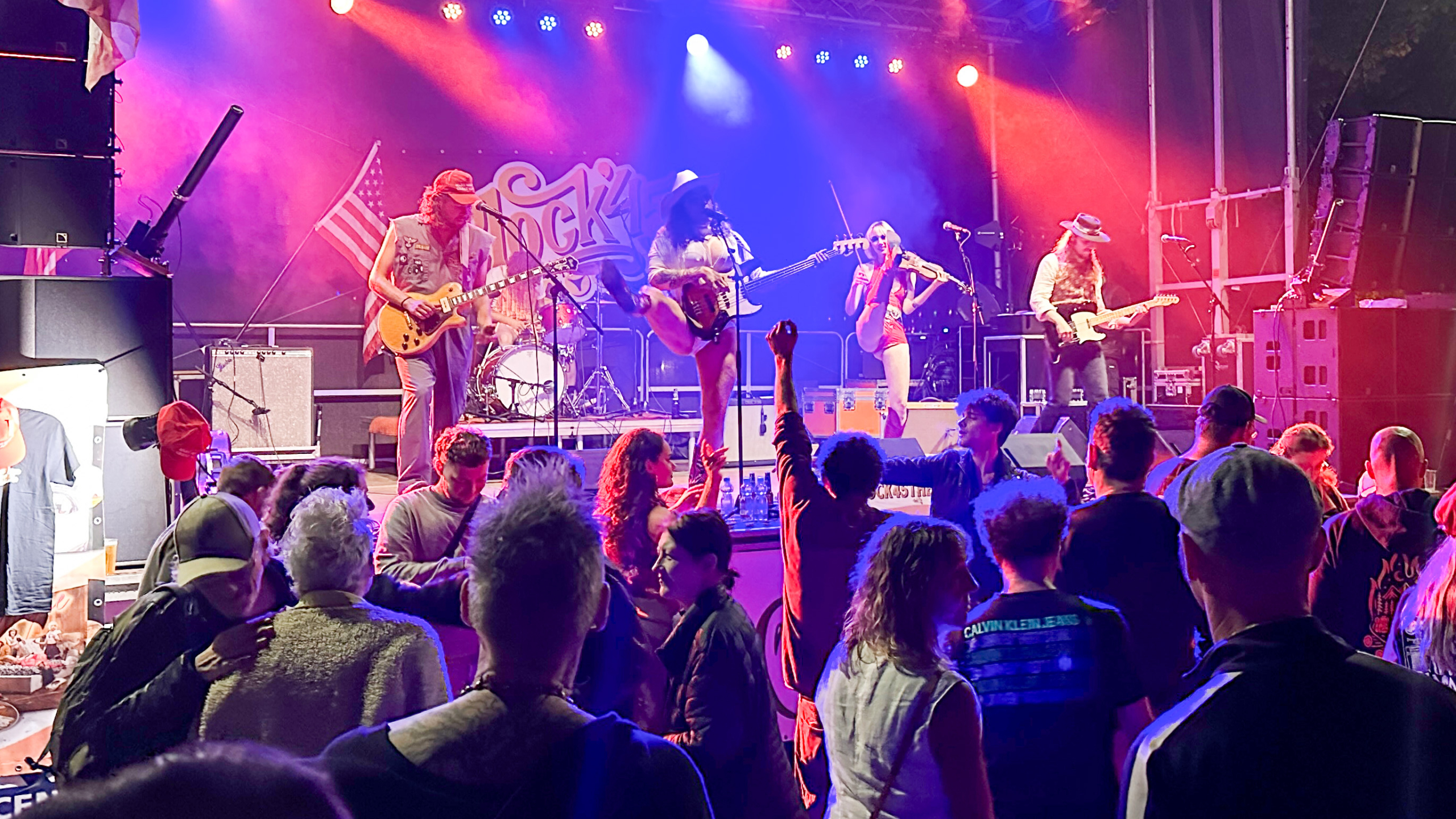 Een live-optreden op een verlicht podium met een band die instrumenten bespeelt en een publiek dat ervoor danst, in een sfeervolle concertomgeving.