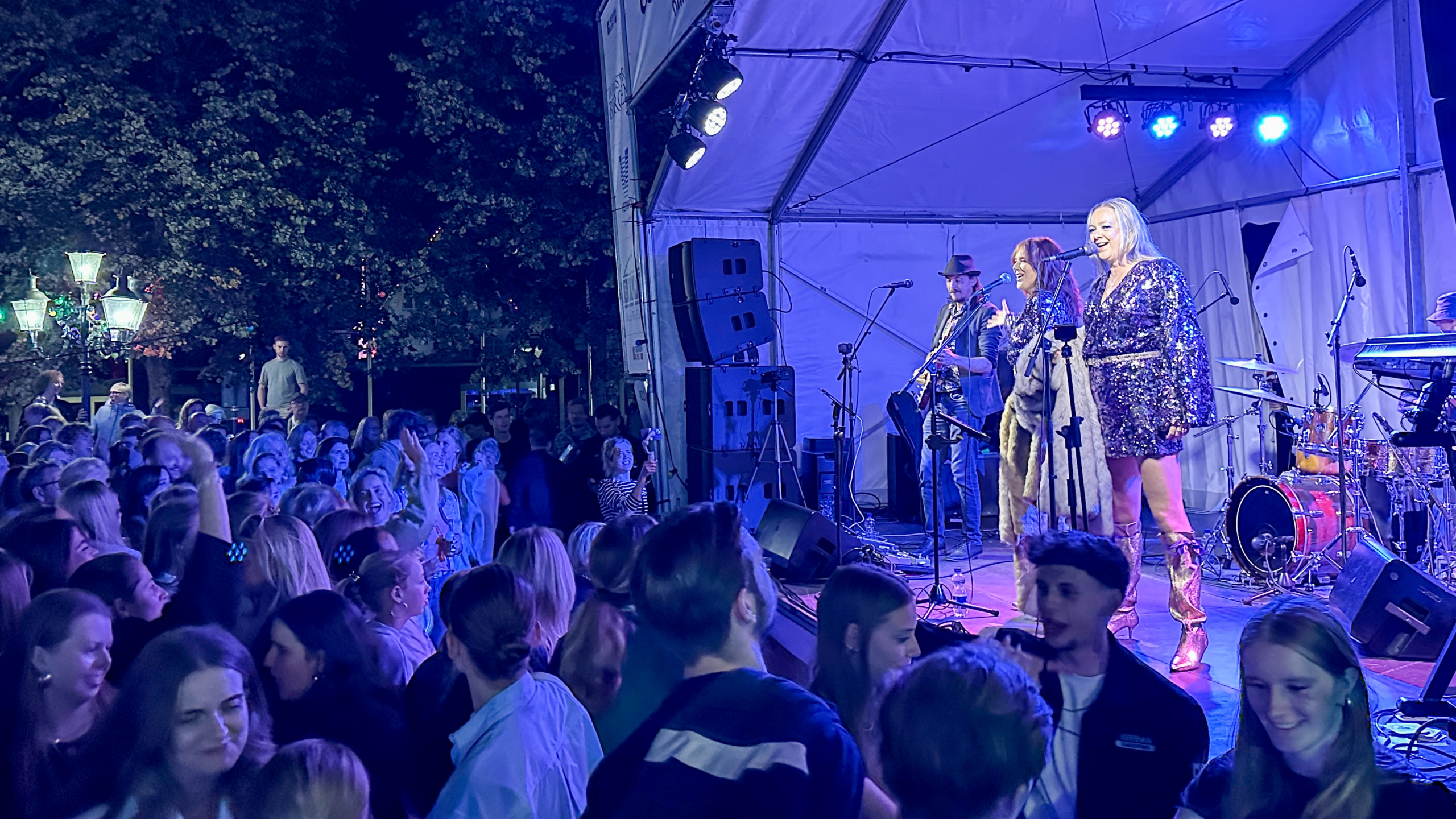 Zangeres in glinsterende outfit en laarzen op een podium, naast een muzikant met gitaar, voor een groot publiek tijdens een openluchtconcert in de avond.