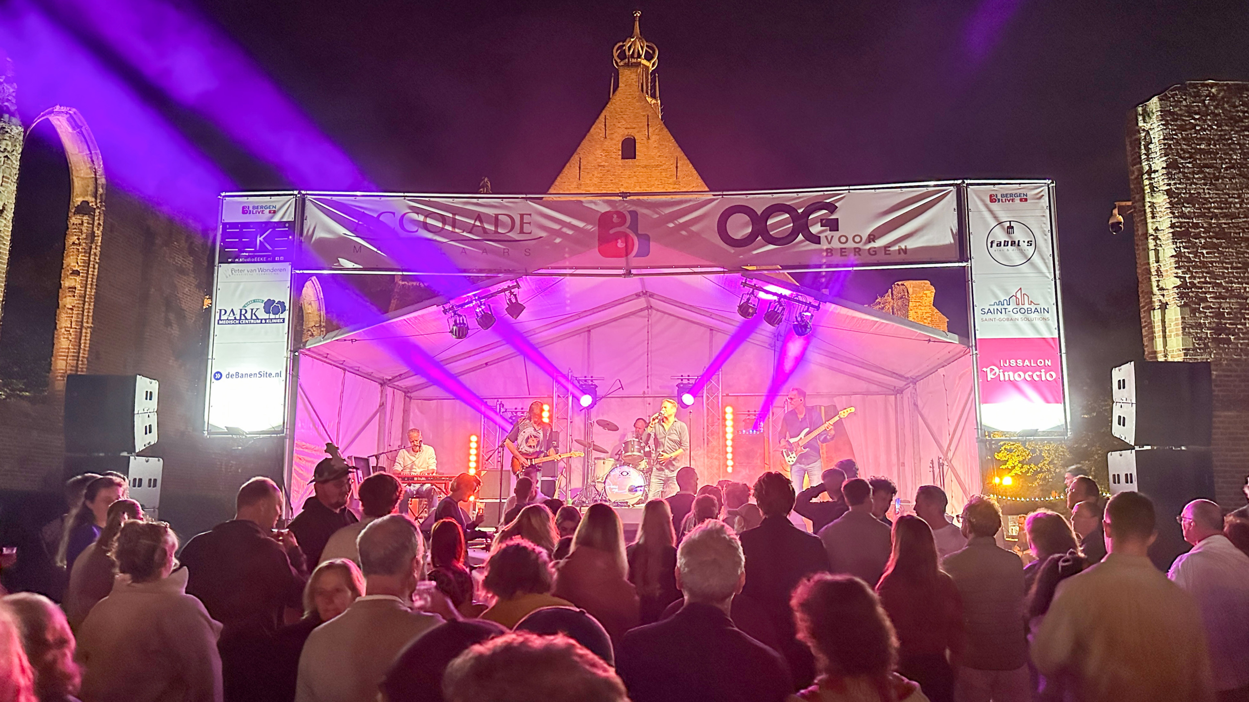 Een nachtelijk openluchtconcert met een band die speelt op een verlichte podiumtent, omringd door een menigte toeschouwers. Het podium is versierd met spandoeken van verschillende bedrijven en heeft gekleurde lichtstralen. Op de achtergrond zijn oude architectonische structuren zichtbaar.