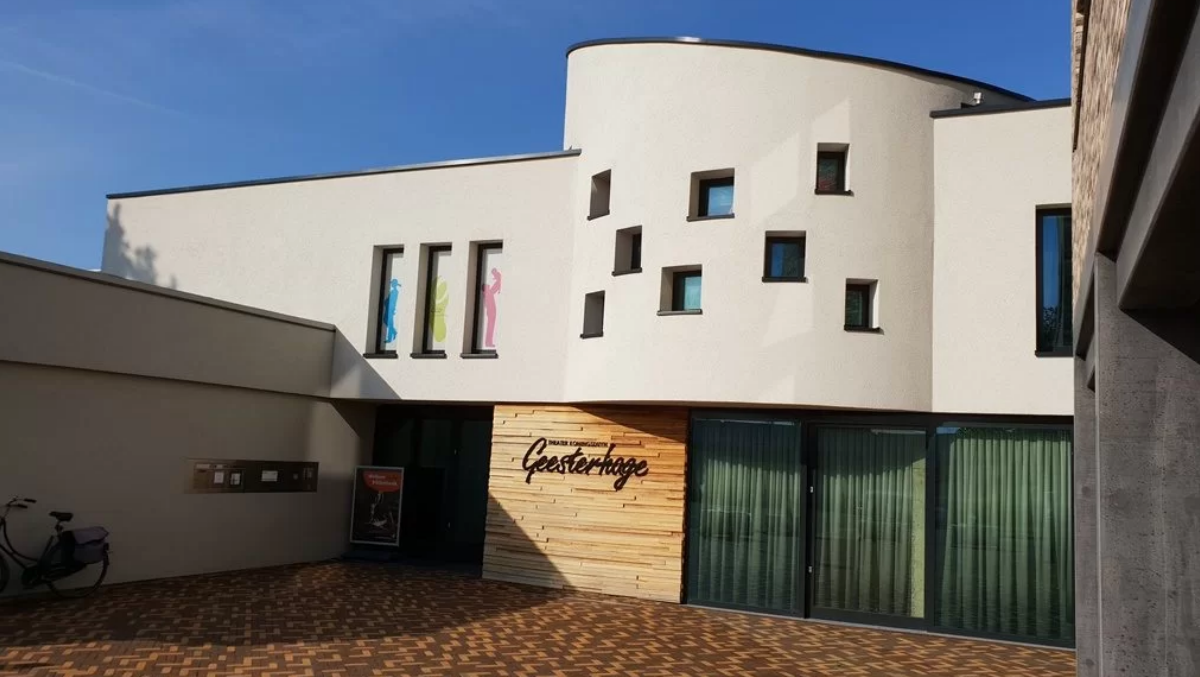 Een modern gebouw met de naam Geesterhage op een houten paneel, voorzien van smalle ramen met silhouetten van figuren in blauw, groen en roze, en een bakstenen vloer op de voorgrond.