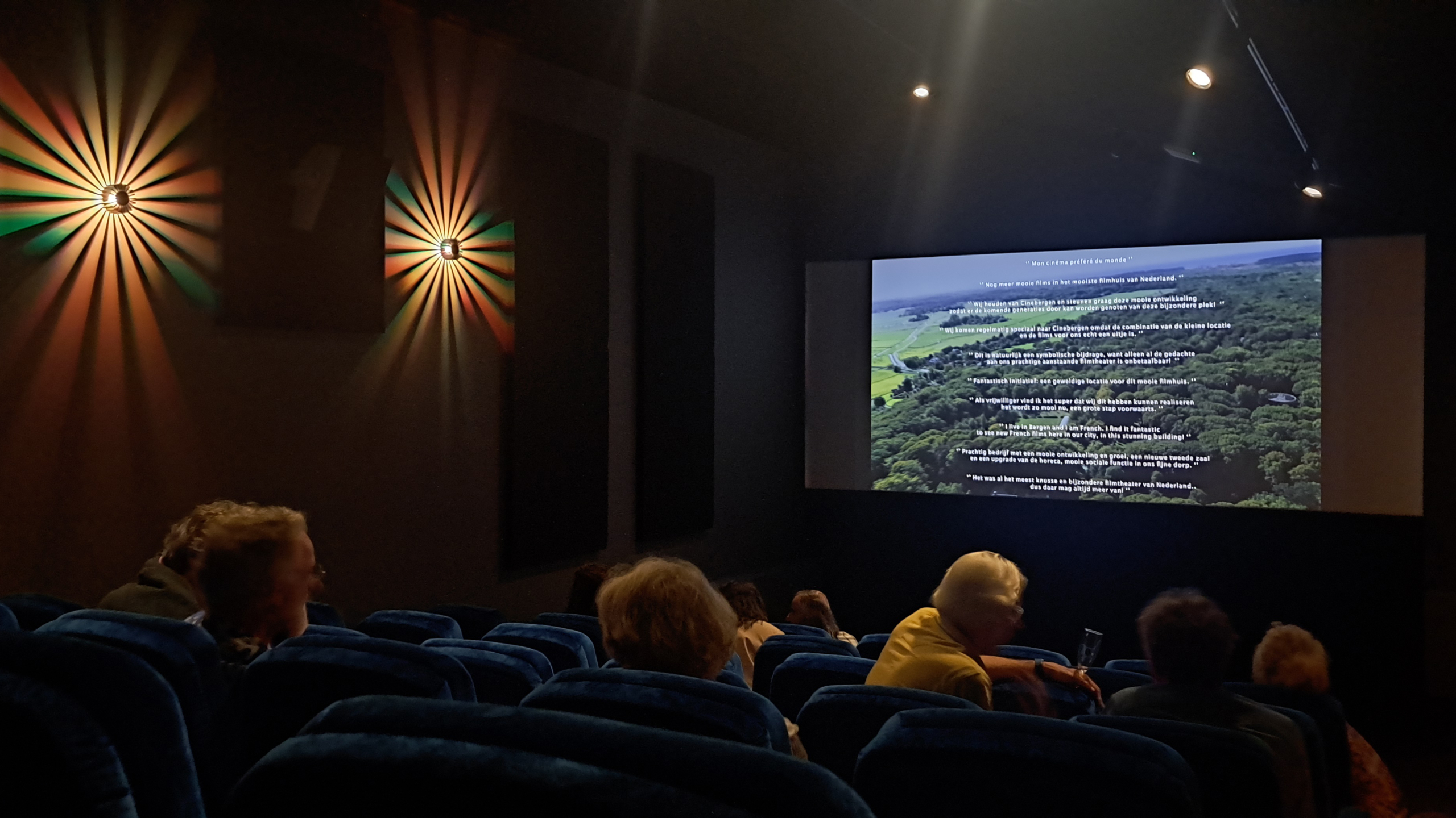 Interieur van een bioscoop met mensen die in blauwe stoelen zitten, met teksten geprojecteerd op een scherm aan de voorkant. Twee kleurrijke muurlampen zijn zichtbaar aan de linkerzijde.