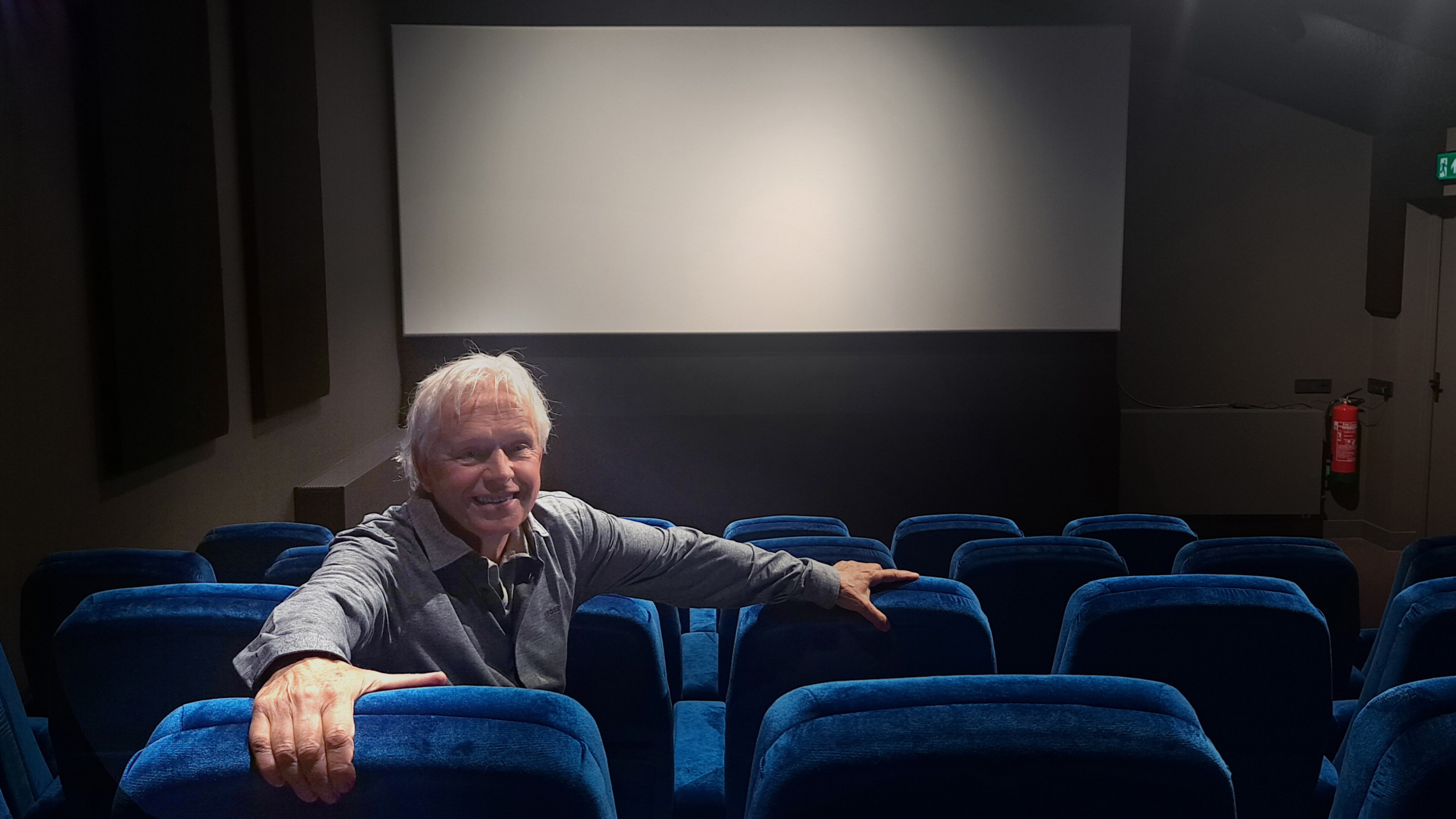 Een oudere man zit lachend in een bijna lege bioscoopzaal met blauwe stoelen, voor een leeg bioscoopscherm.