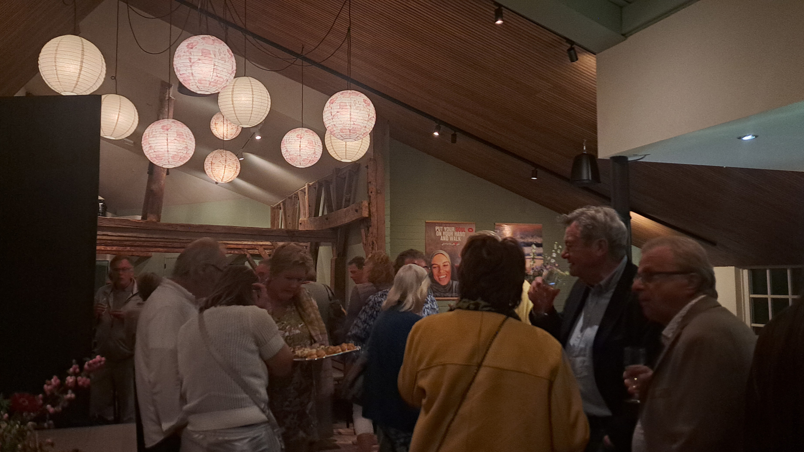 Groep mensen in een sfeervolle ruimte met ronde, papieren lampen aan het plafond; sommigen hebben een drankje vast, terwijl anderen met elkaar in gesprek zijn.