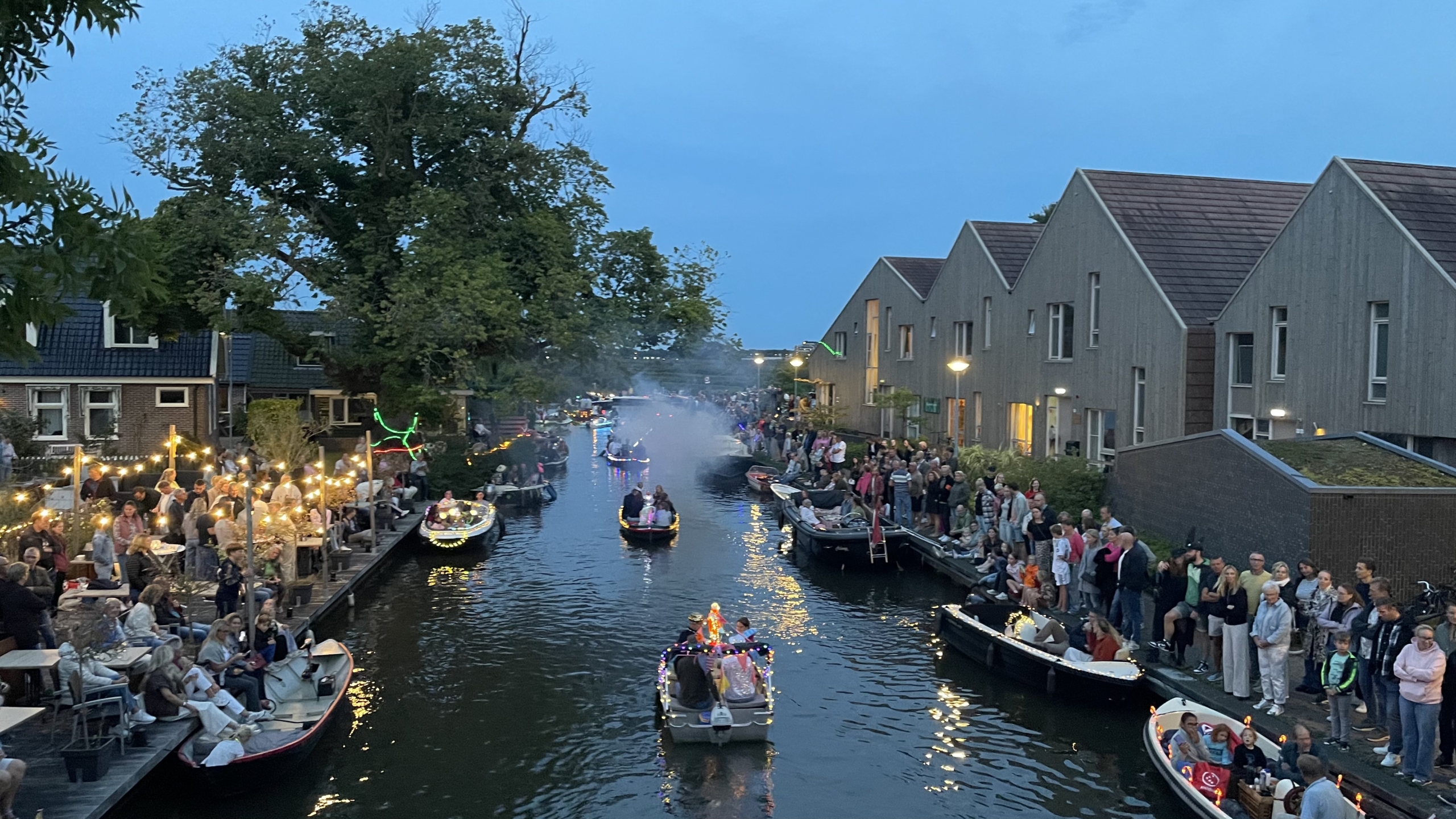 Avondbeeld van een kanaal in Nederland met versierde boten die langs drijven tijdens een evenement; toeschouwers staan langs beide oevers, sommige boten zijn versierd met lichtjes en mensen kijken toe.