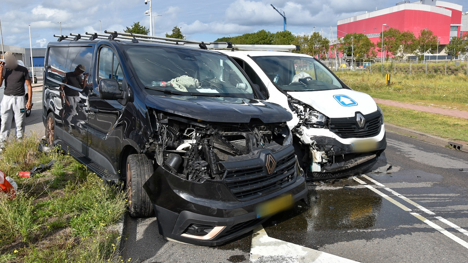 Twee bestelwagens met aanzienlijke schade aan de voorkant na een botsing, geparkeerd aan de kant van de weg met vloeistof op het wegdek. Er staat een man naast het zwarte busje.
