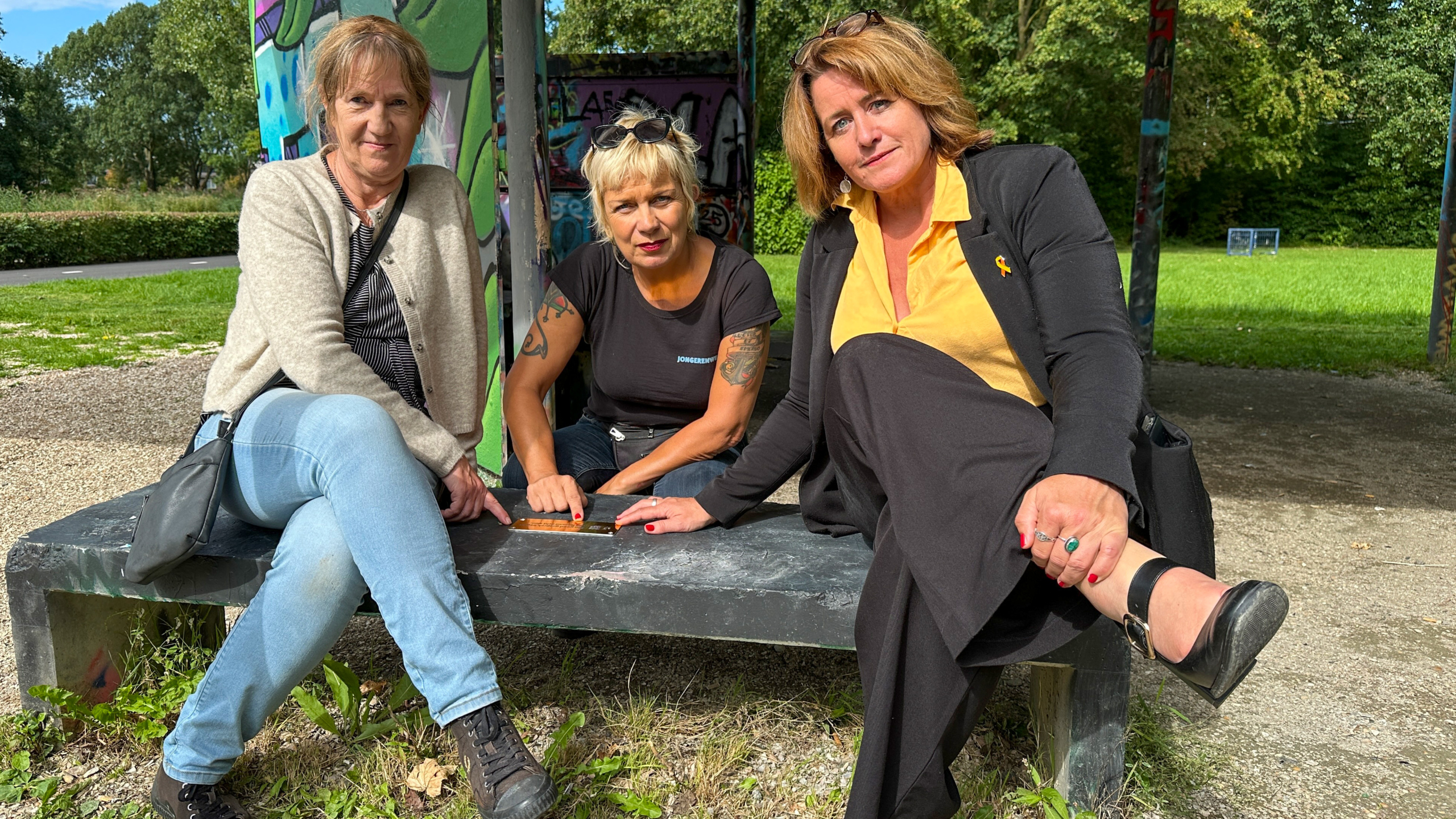 Drie vrouwen zitten buiten op een bankje, omringd door groen en graffiti, terwijl ze een klein bord bekijken. De vrouw links draagt een beige vest, de vrouw in het midden heeft een zwarte T-shirt en tatoeages op haar armen, en de vrouw rechts draagt een geel shirt met een zwart jasje.