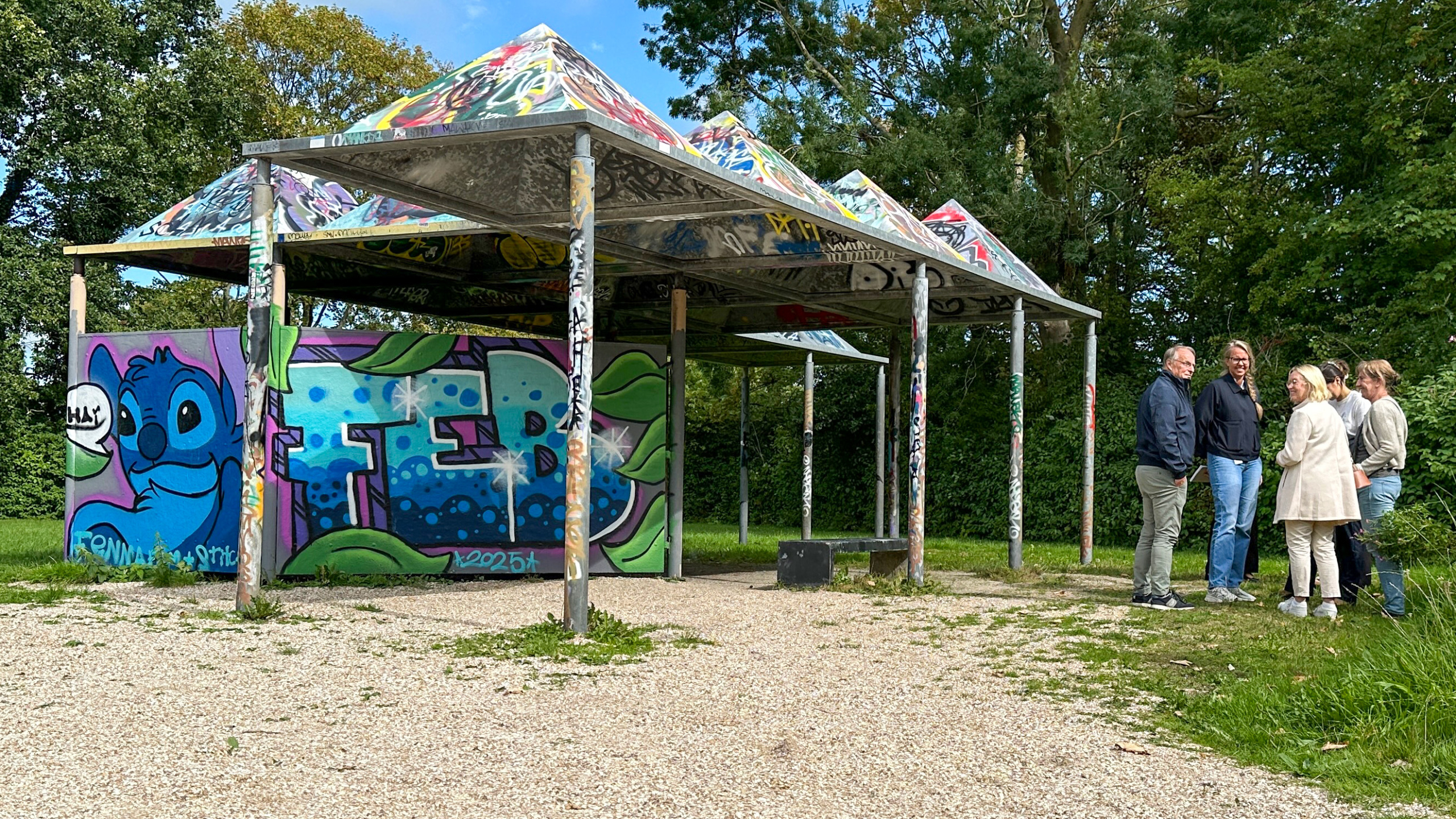 Een groep mensen staat naast een beschilderd kunstwerk met graffiti onder een met graffiti bedekt afdak in een parkachtige omgeving, omringd door bomen.