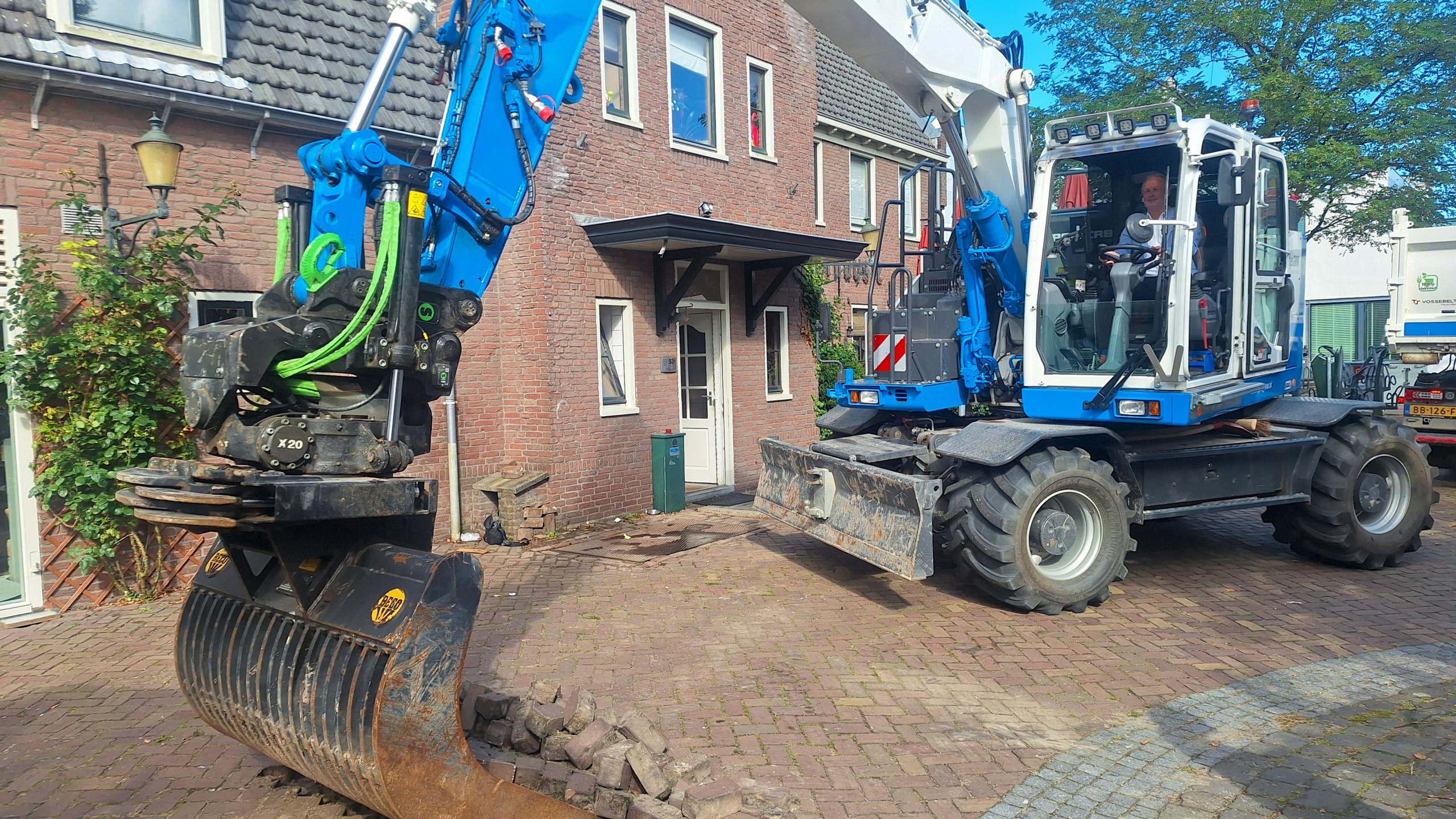 Een graafmachine met blauwe en witte details graaft stenen op een straat voor een bakstenen gebouw. Een persoon zit in de cabine van de machine, en op de achtergrond is een vrachtwagen zichtbaar. Het is een zonnige dag.