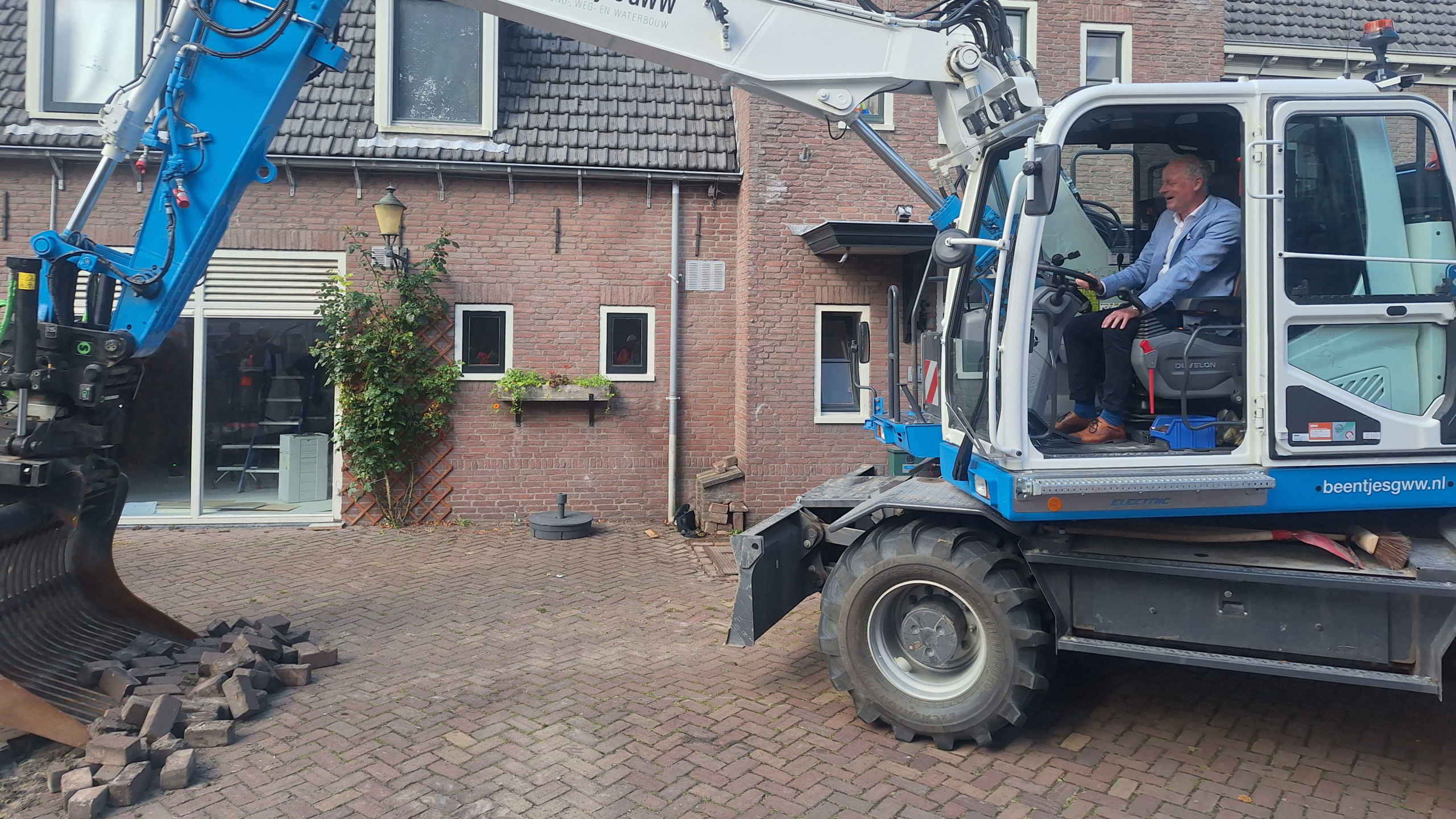 Een man bestuurt een blauwe graafmachine voor een bakstenen huis met gebogen dakpannen, terwijl hij stenen op een verharde binnenplaats verplaatst.