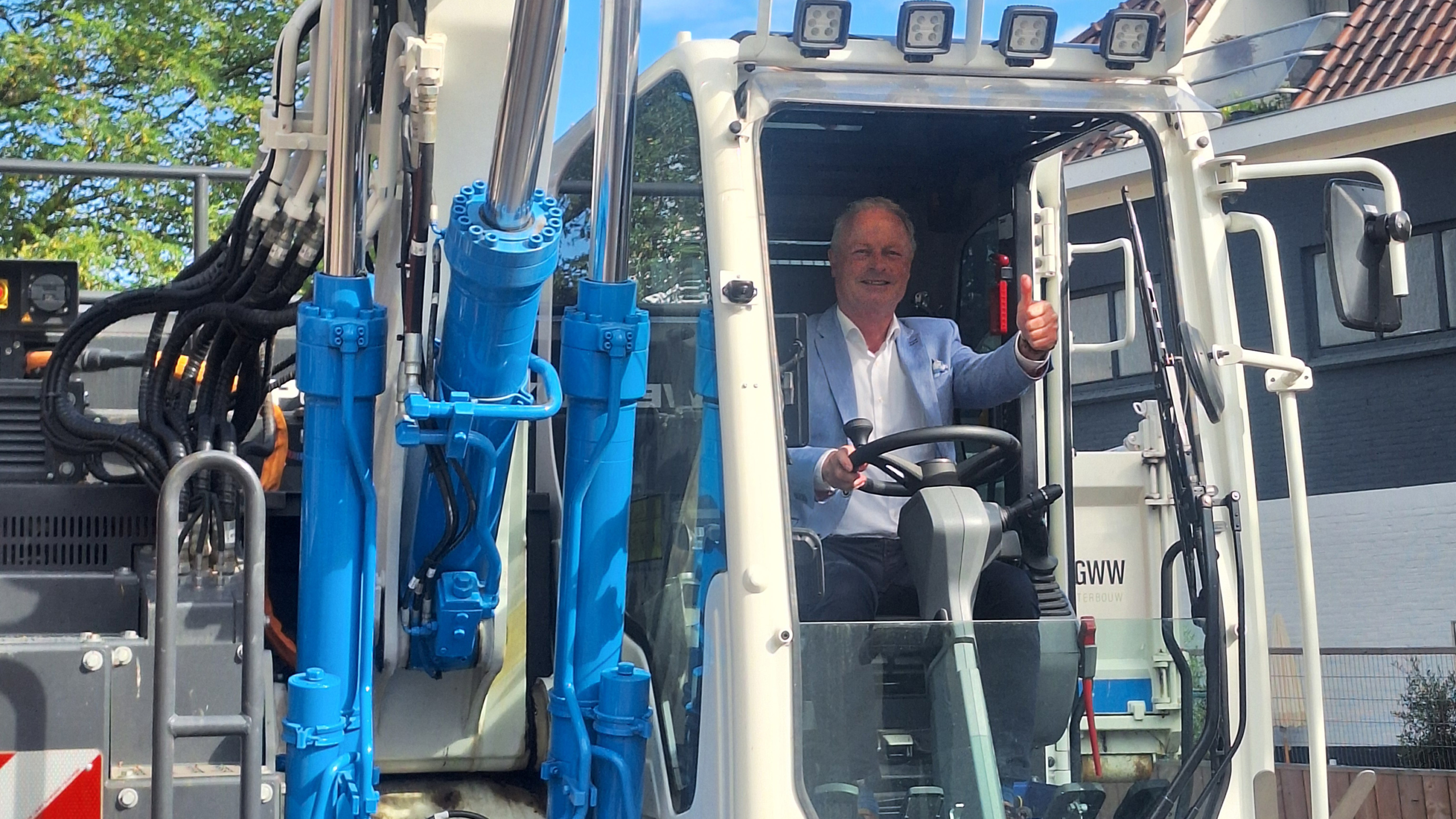 Een man in een lichtblauw pak geeft een duim omhoog terwijl hij in de cabine van een zware bouwmachine zit, omgeven door blauwe hydraulische onderdelen en met een gebouw op de achtergrond.