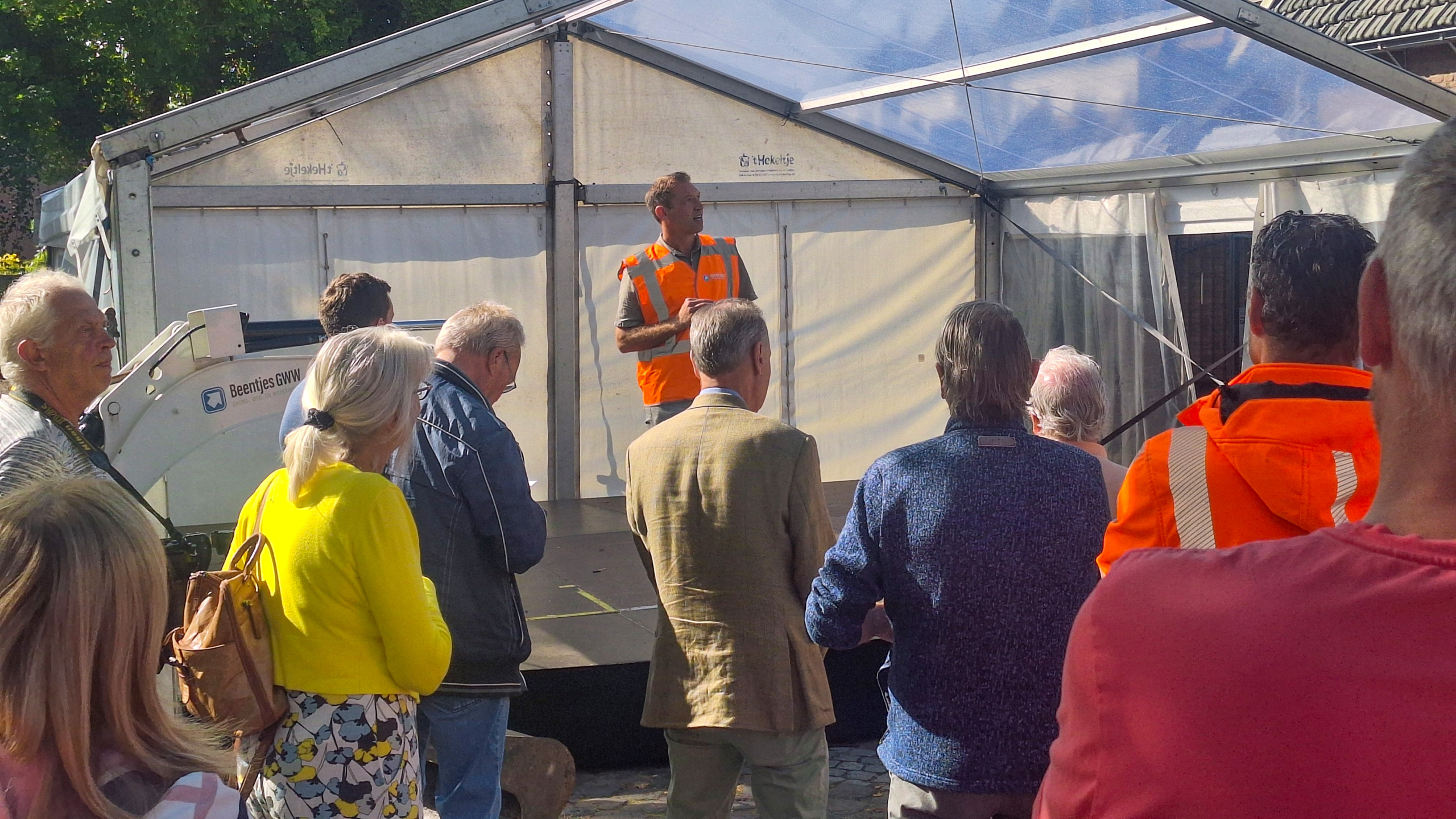 Een groep mensen luistert naar een spreker in een oranje veiligheidsvest onder een tentconstructie in de buitenlucht.