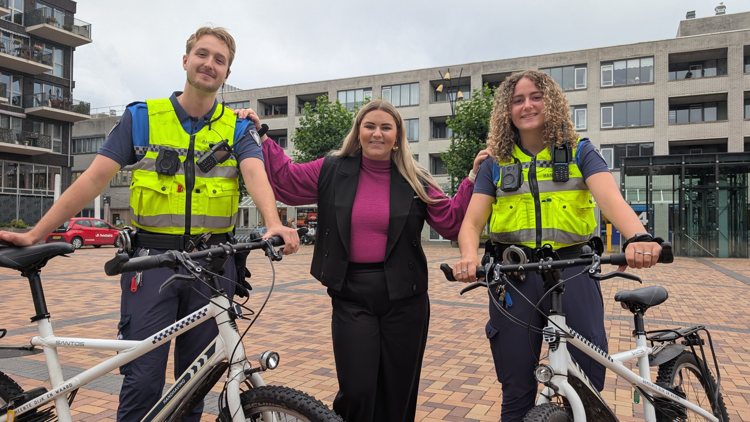 Twee handhavers in uniform staan naast een vrouw, allemaal glimlachend, met fietsen op de voorgrond en een stedelijke omgeving als achtergrond.