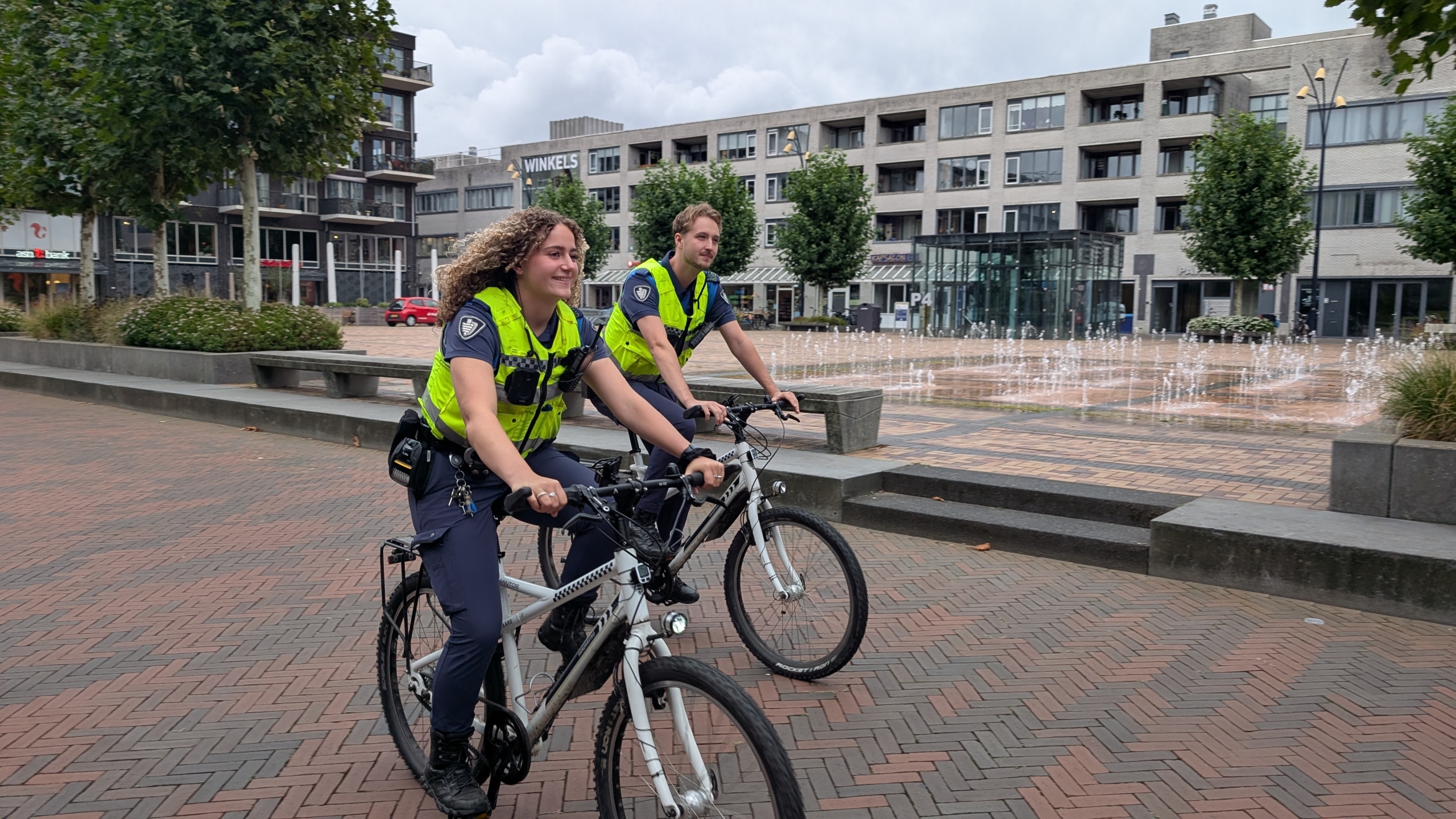 Twee politieagenten fietsen op mountainbikes over een plein met een fontein op de achtergrond. Ze dragen gele veiligheidsvesten en zijn in uniform. Het plein is omringd door bomen en moderne gebouwen.