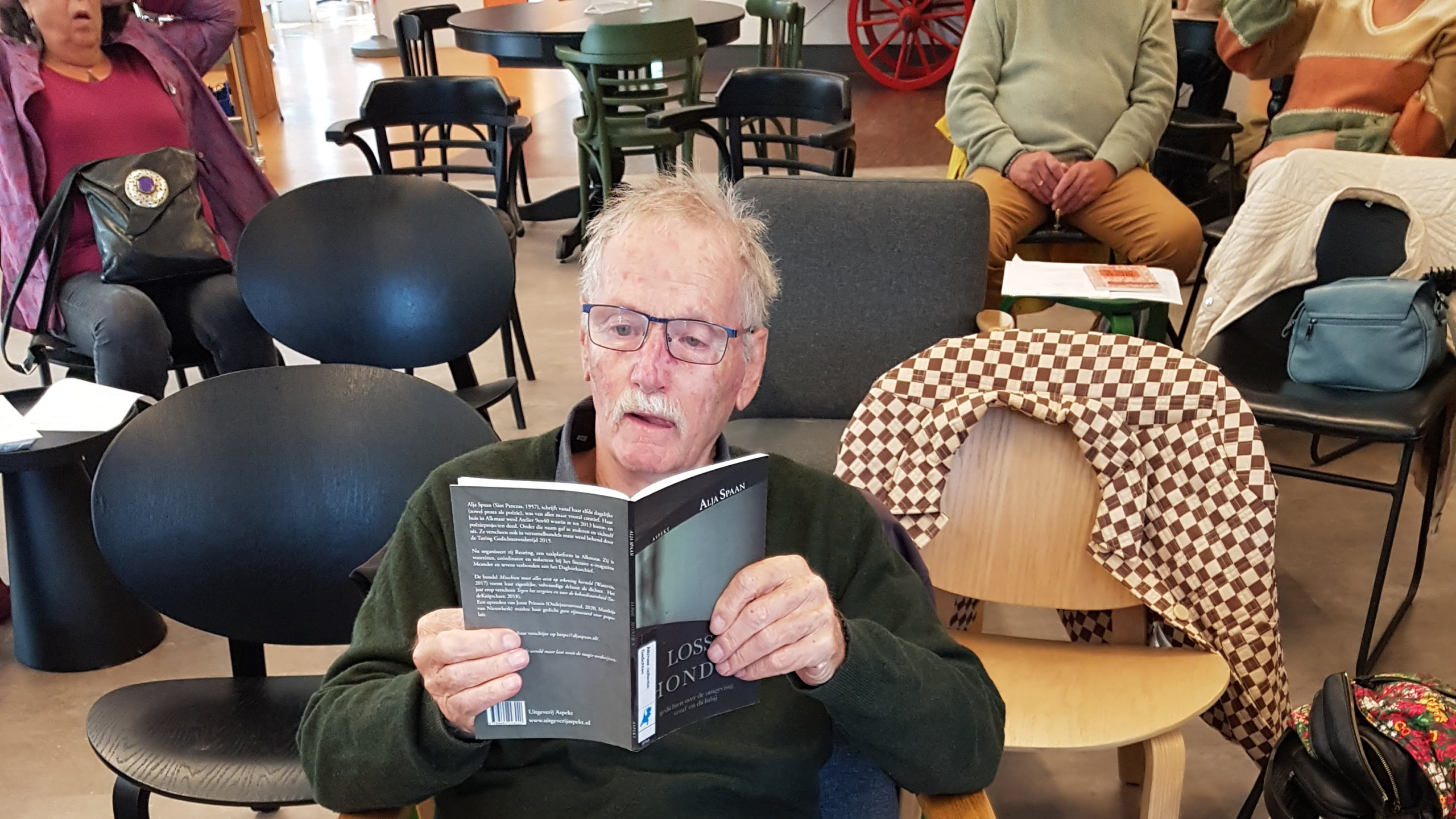 Een oudere man leest een boek in een ruimte met andere mensen, zittend op stoelen. Sommige mensen lijken aandachtig te luisteren, terwijl anderen zich ontspannen of praten. De omgeving is binnen, met verschillende soorten stoelen en tafels.