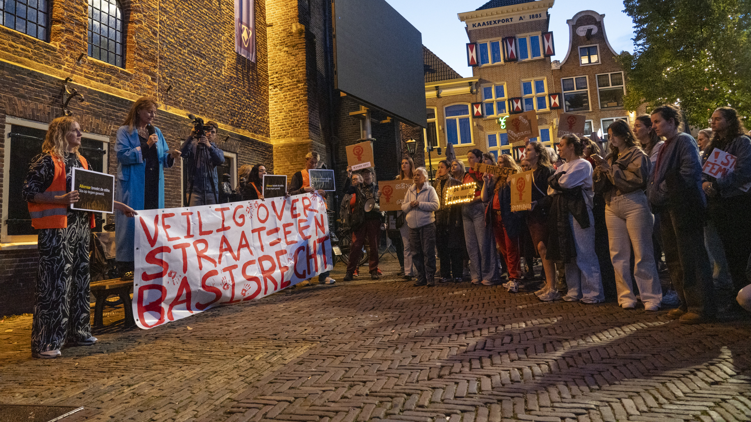 Een groep mensen staat buiten bij een demonstratie met borden en spandoeken die pleiten voor veilige straten en basisrechten, gefotografeerd in de schemering voor een bakstenen gebouw.