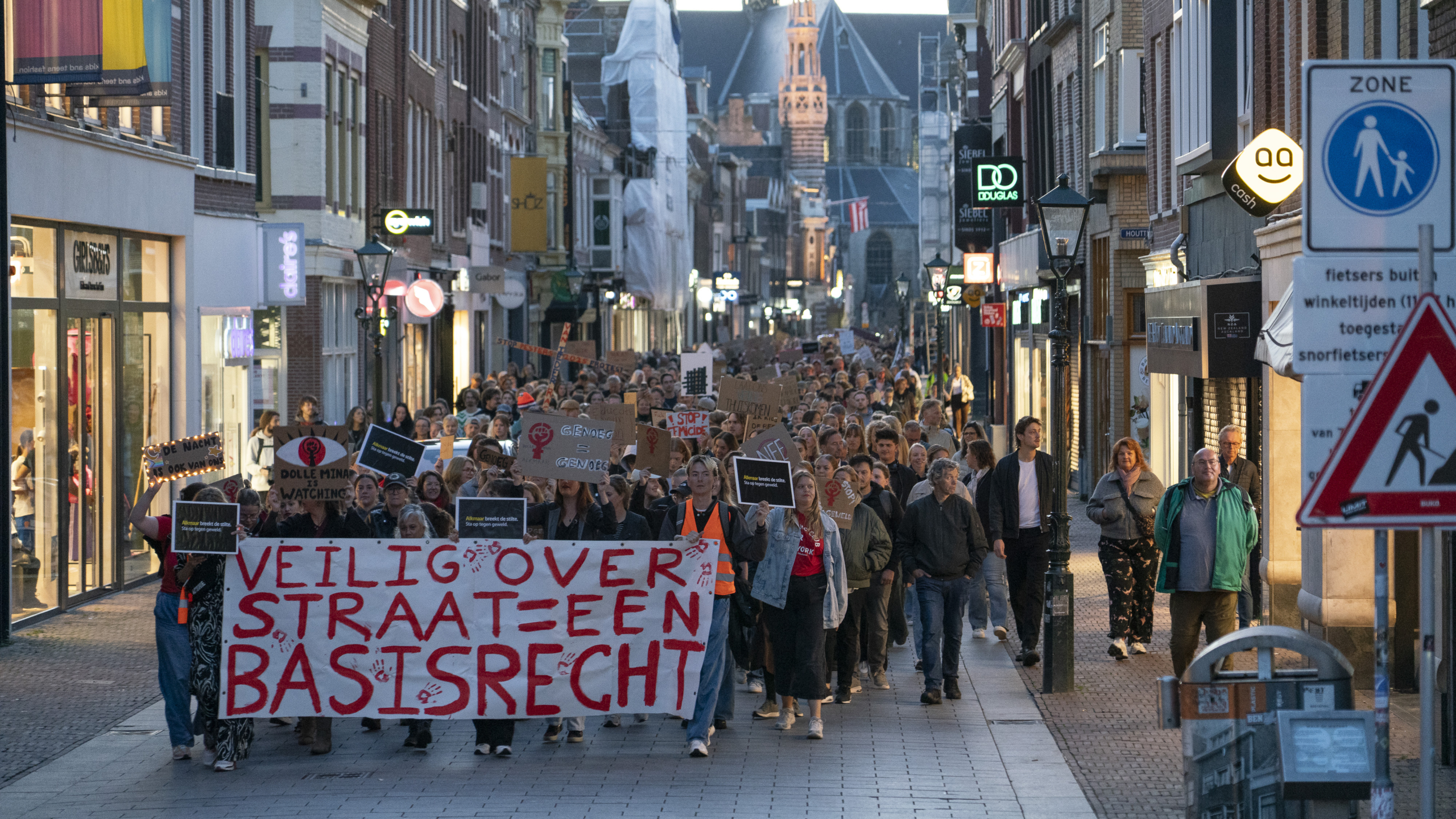 Mensen lopen in een protestmars door een winkelstraat met spandoeken waarop teksten staan als "Veilig over straat = een basisrecht". Op de achtergrond zijn historische gebouwen zichtbaar.