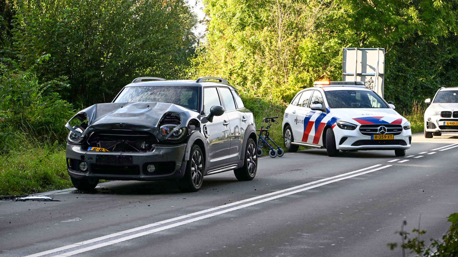 Auto met aanzienlijke schade aan de voorkant staat naast een politieauto op een weg, met een rolator op de achtergrond bij de bosrand.