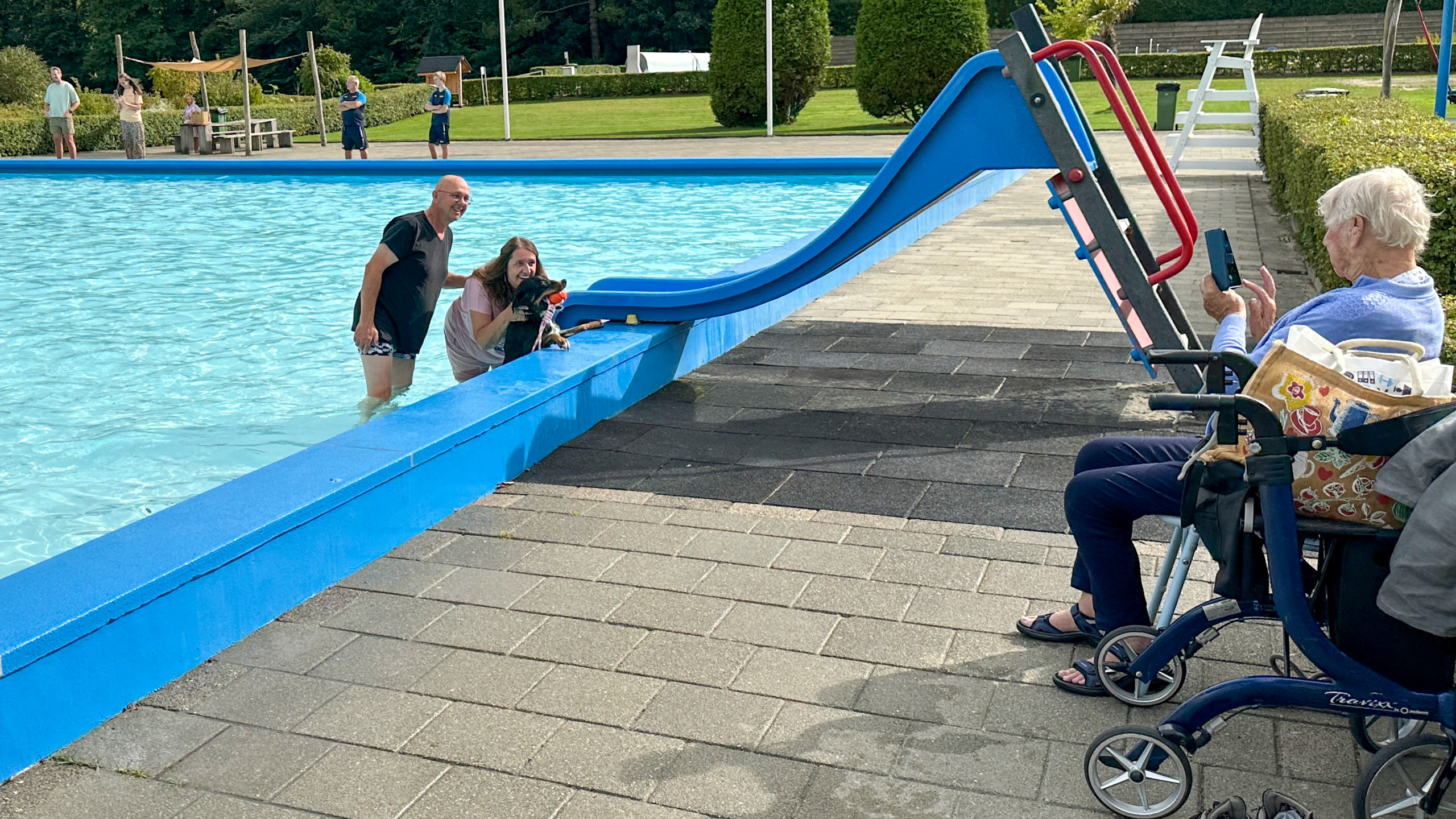 Twee mensen en een hond in een zwembad naast een blauwe glijbaan, met een oudere vrouw die op een rollator zit en een foto maakt.