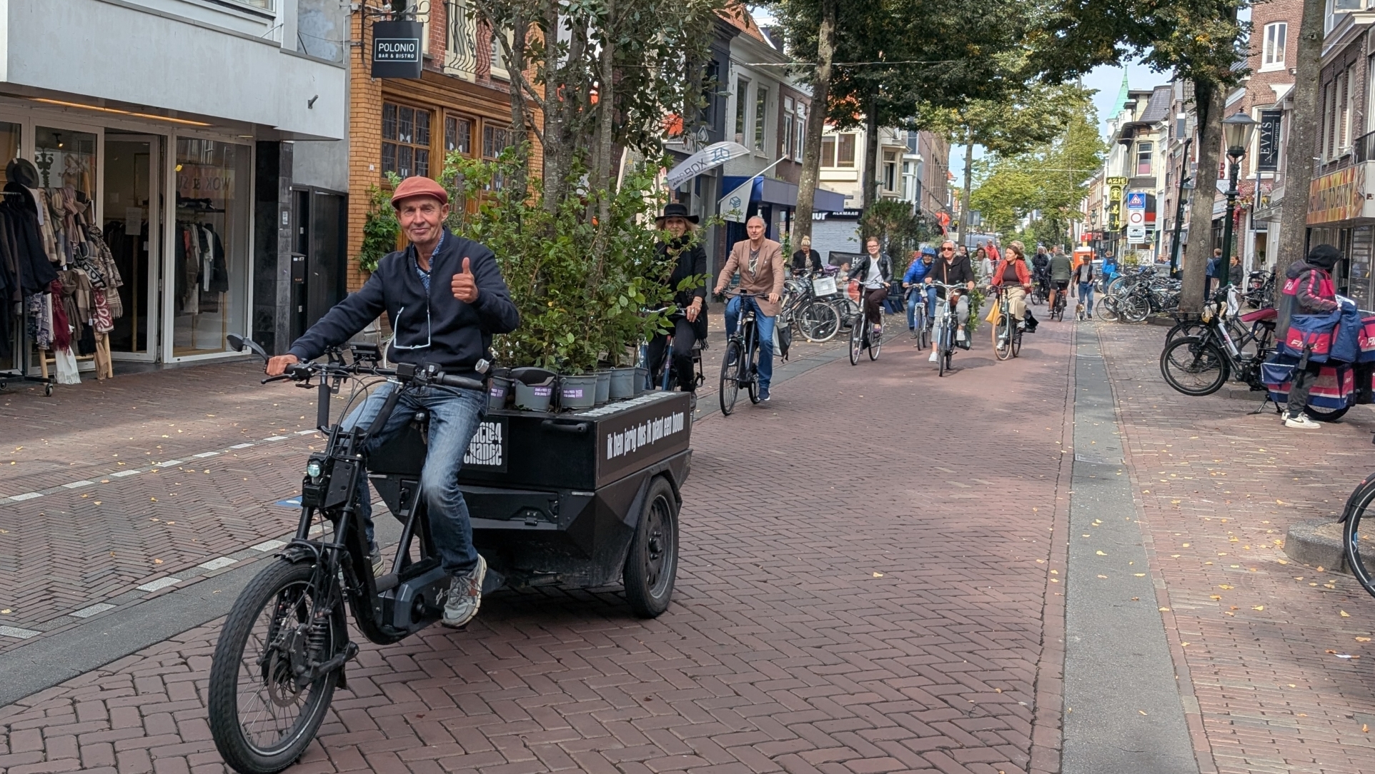 Een man rijdt op een bakfiets door een drukke winkelstraat vol fietsers in Nederland; hij draagt een pet en steekt zijn duim op, terwijl hij een lading planten vervoert.