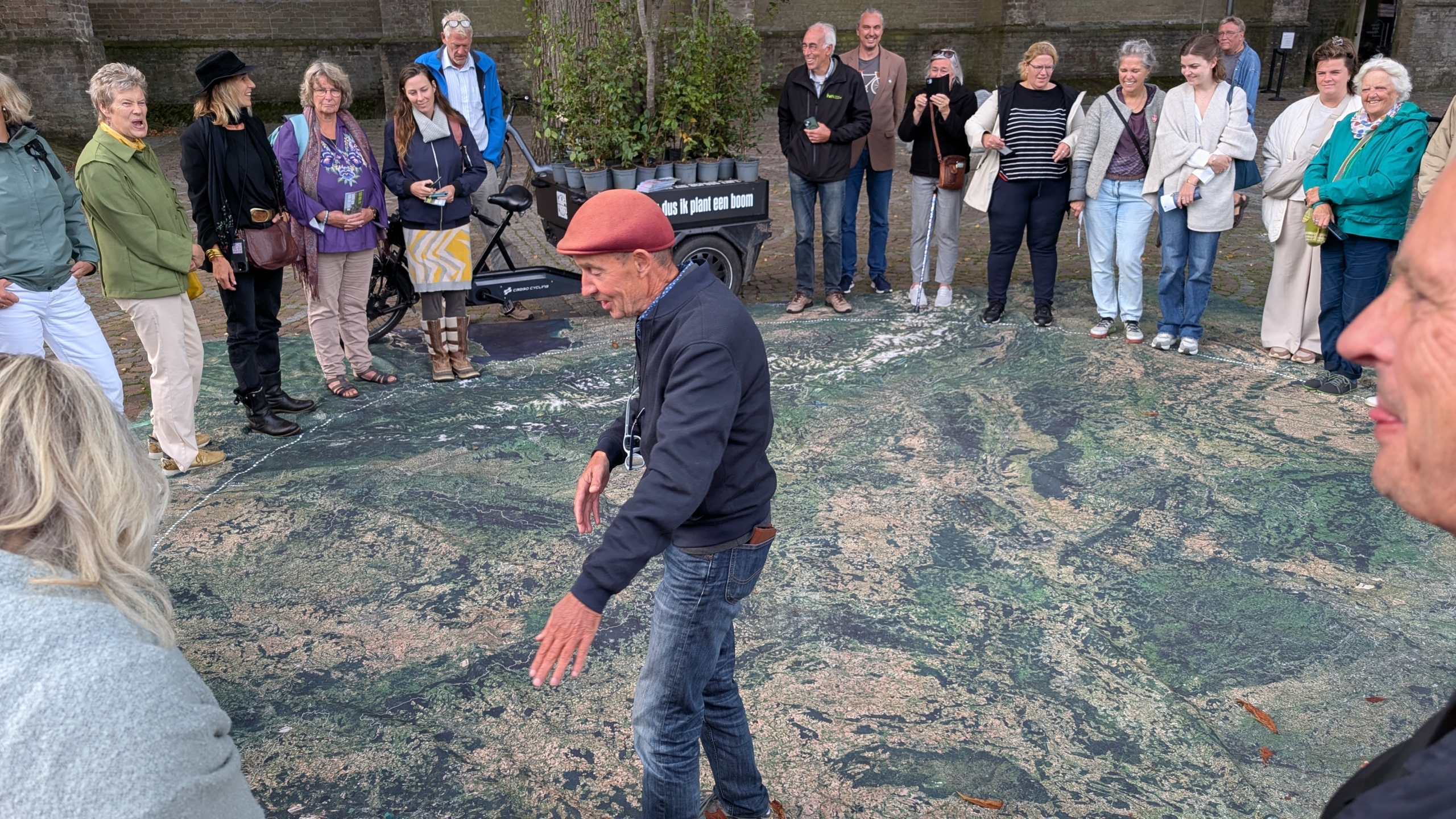 Een groep mensen staat in een cirkel om een man die op een grote kaart op de grond loopt, buiten in een open ruimte.