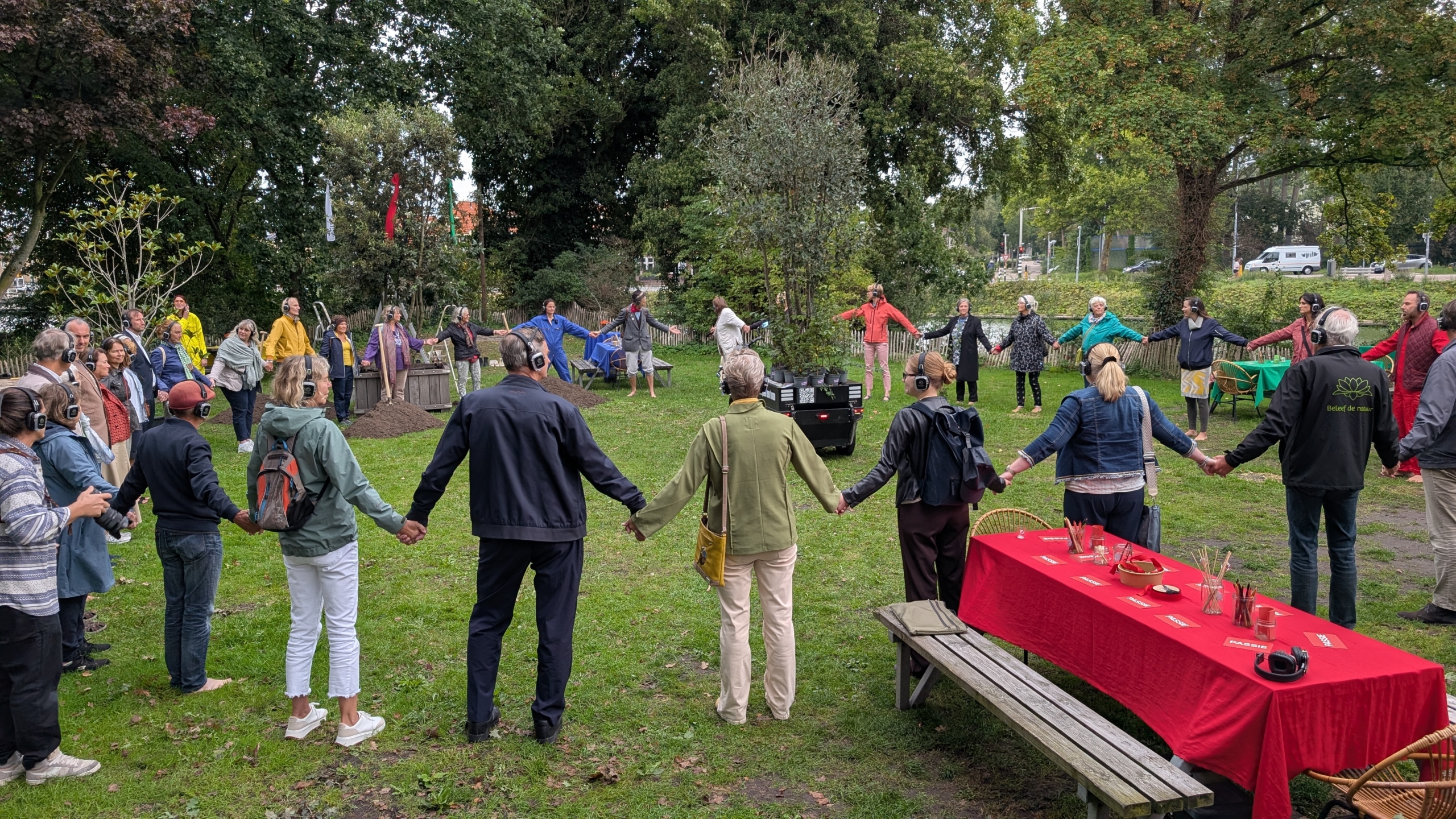 Mensen vormen een cirkel in een bosrijke omgeving, waarbij ze elkaars handen vasthouden en koptelefoons dragen, naast een tafel bedekt met een rood tafelkleed.