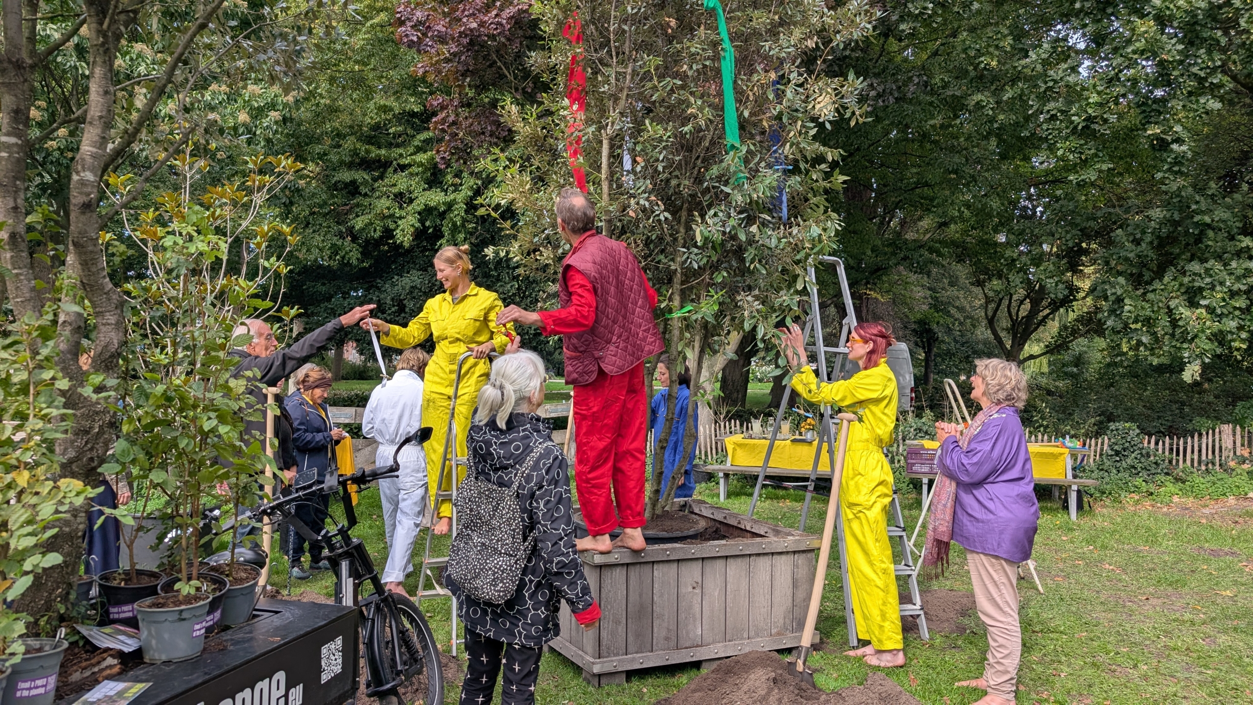 Groep mensen in kleurrijke kleding die linten in een boom hangen in een parkachtige omgeving.