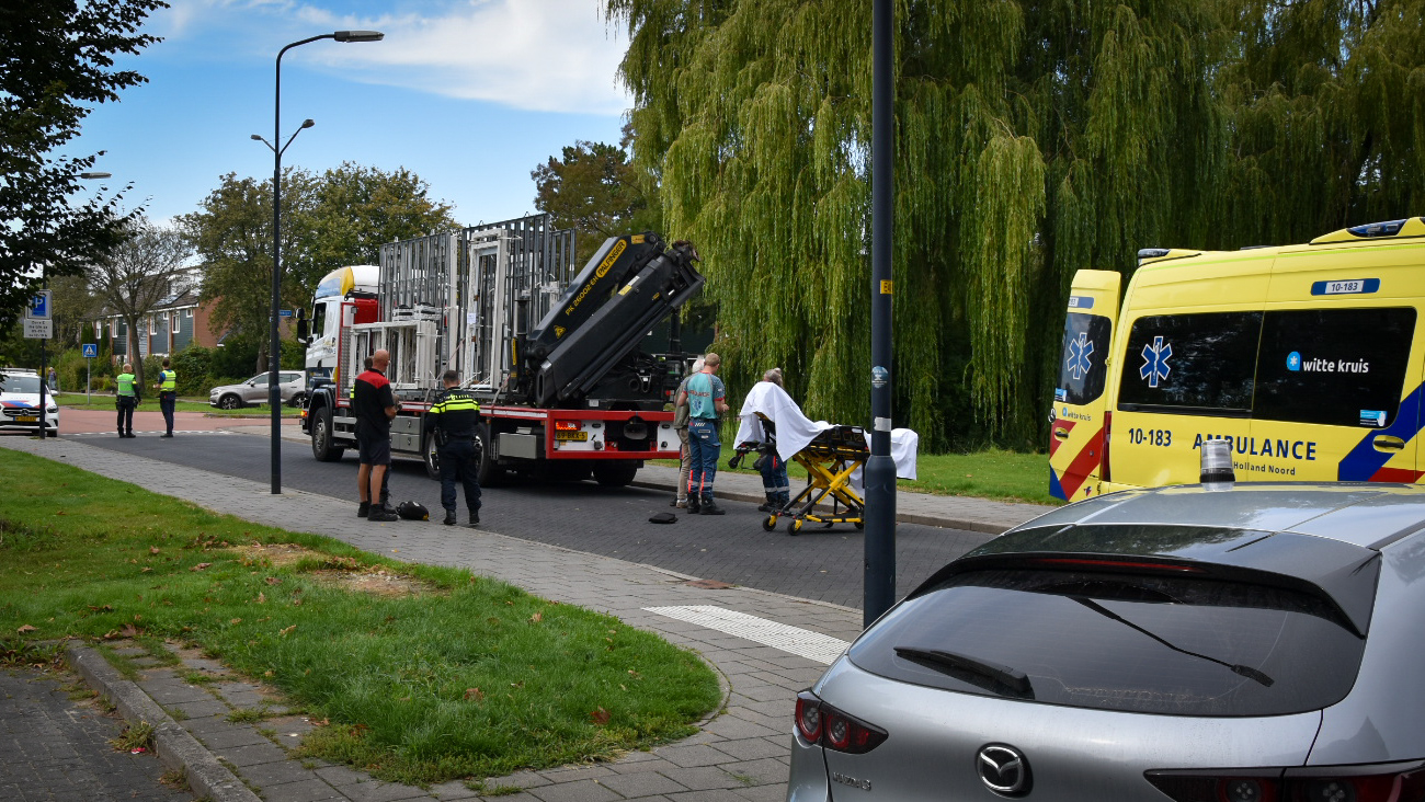 Een verkeersincident op straat met een ambulance, politieagenten, en een vrachtwagen. Een groepje mensen staat rondom een brancard naast de vrachtwagen.