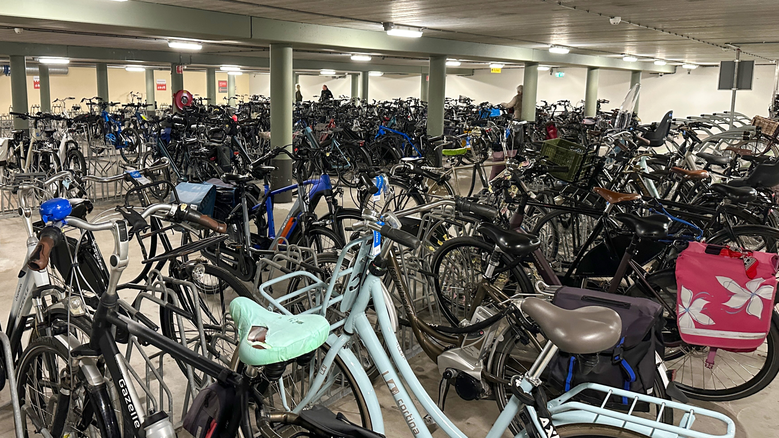 Overvolle fietsenstalling in een parkeergarage met rijen fietsen dicht op elkaar, waaronder een blauwe Cortina fiets met een mintgroen fietszitje en een fiets met een roze fietstas.