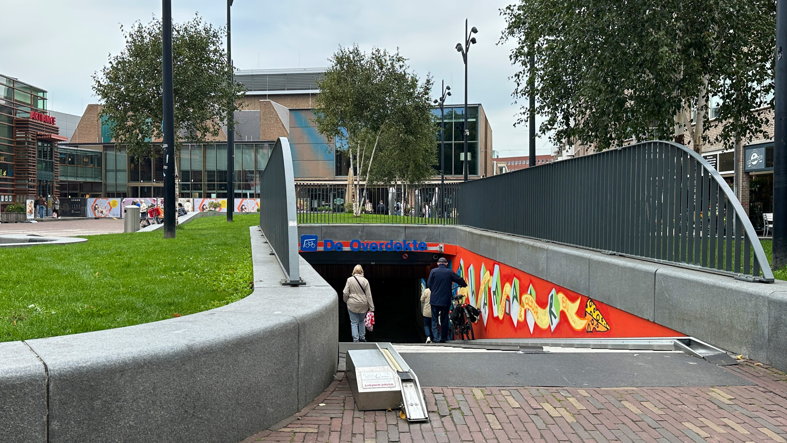 Ingang van een fietstunnel genaamd 'De Overdekte' met een kleurrijke muurschildering, omgeven door een groen grasveld en moderne gebouwen op de achtergrond.