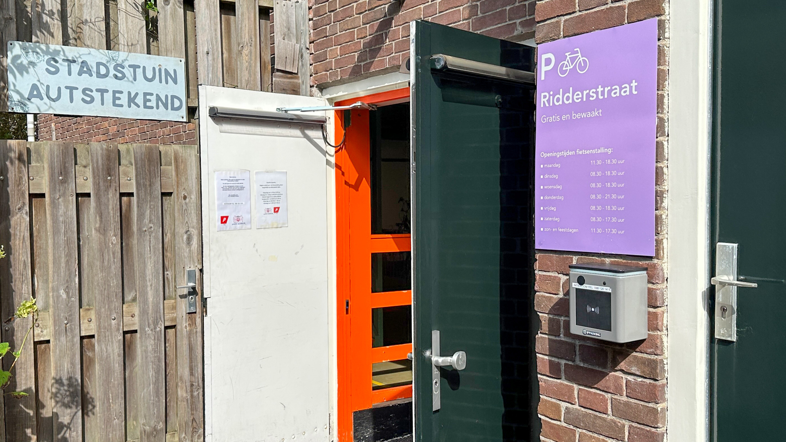Ingang van Stadstuin AuteStekend, met een open groene deur en een paarse poster met openingstijden van de fietsenstalling Ridderstraat.