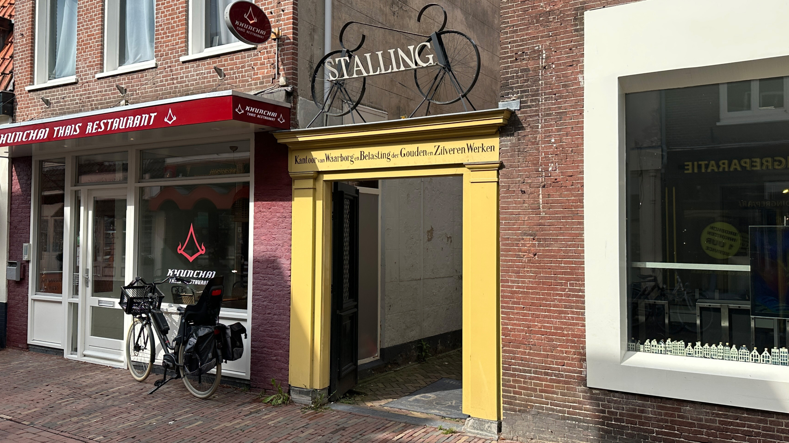 Een Thais restaurant genaamd Khunchai met een fiets geparkeerd voor de deur en een gele doorgang naar een fietsstalling aan de rechterkant van een straat in een stedelijke omgeving.