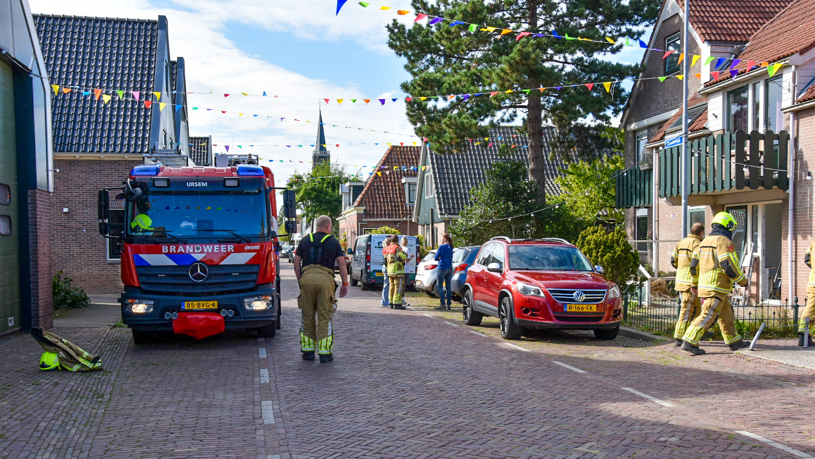 Brandweerwagen en brandweerlieden in een woonstraat met vlaggetjes, naast geparkeerde auto's en een rijtjeshuis.