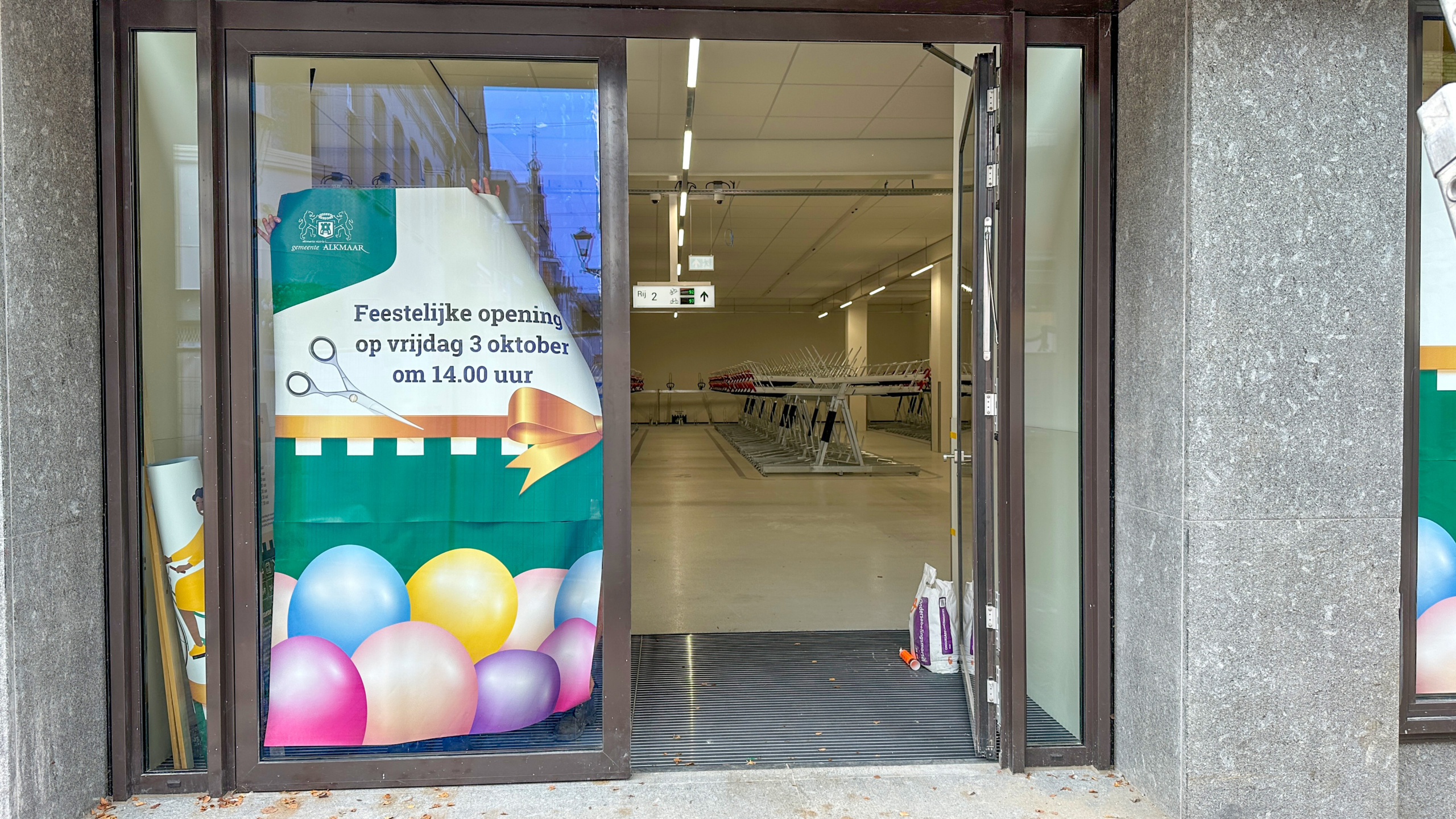 Een openstaande deur naar een lege winkelruimte met een poster op het raam die de feestelijke opening op vrijdag 3 oktober om 14:00 uur aankondigt, versierd met ballonnen en een afbeelding van een schaar en een strik.
