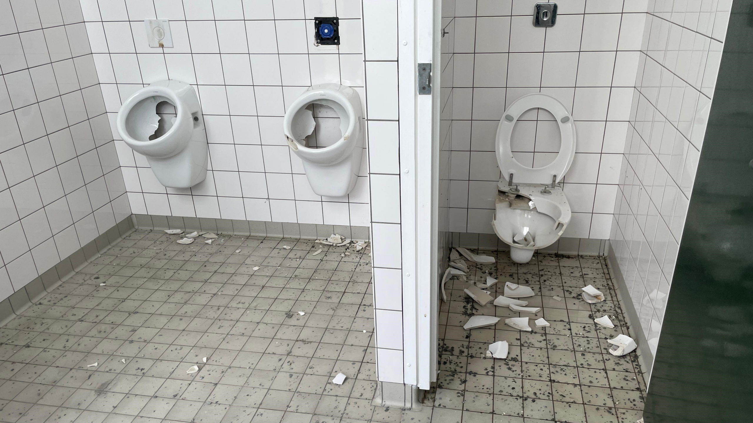 Vernielde sanitaire voorzieningen in een badkamer met kapotte urinoirs en een toilet, omgeven door witte tegels en verspreid puin op de vloer.