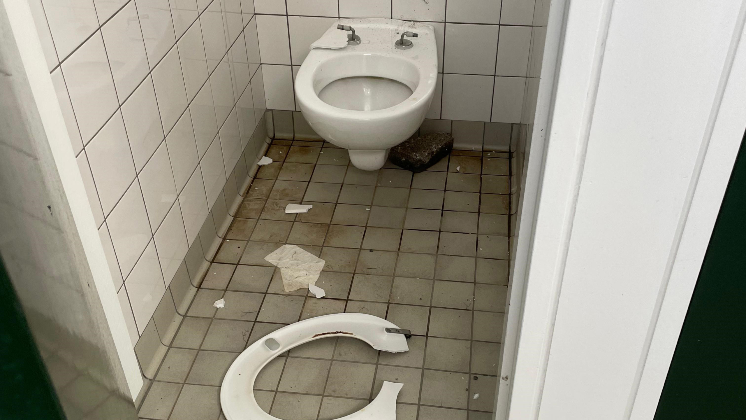 Een toilet in een vervallen staat met een gebroken toiletring op de vloer en vieze tegels en muren.
