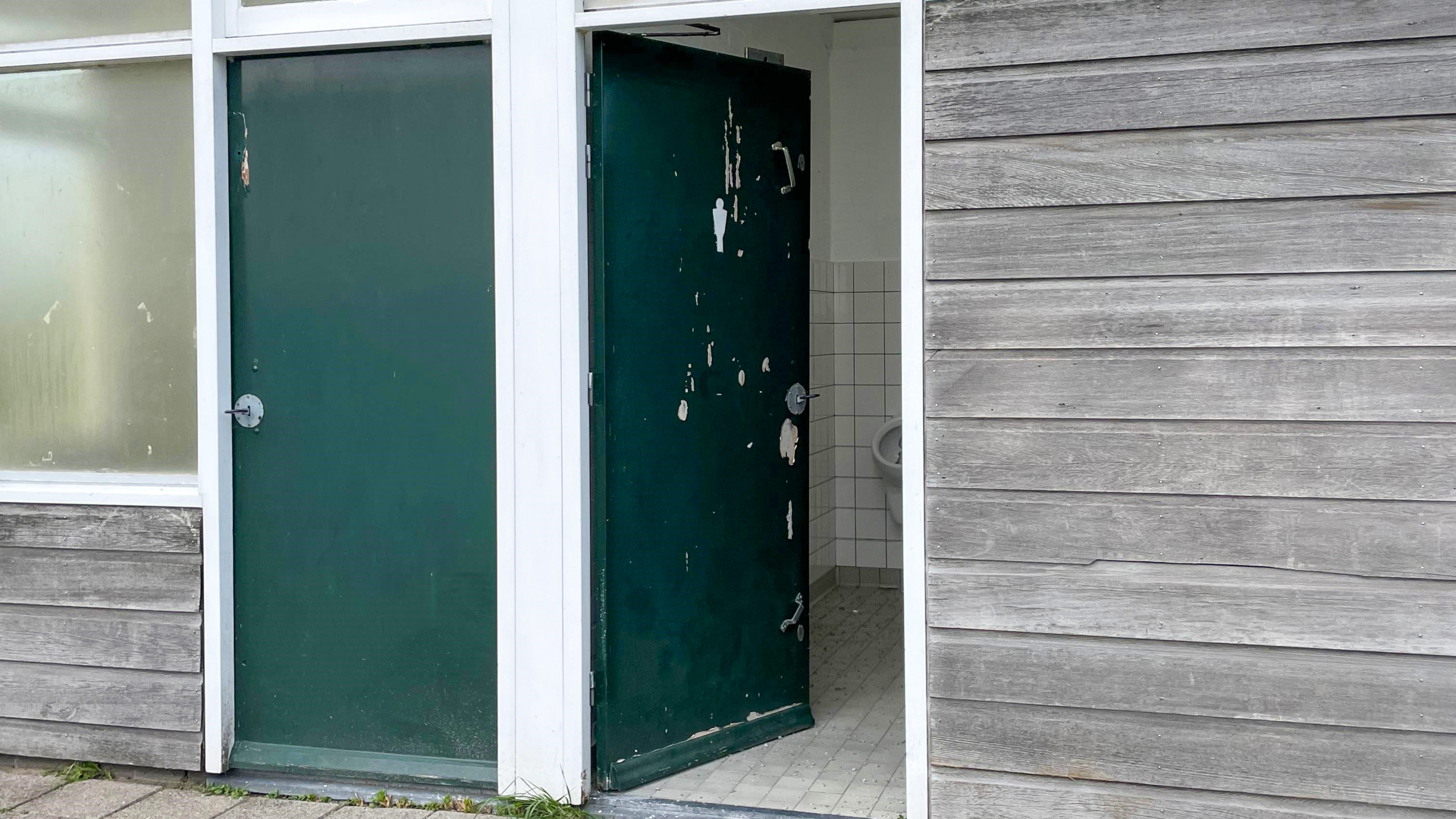 Een groen, afgebladderd deur van een openbaar toilet met tegelvloer, deels geopend in een houten gebouw.