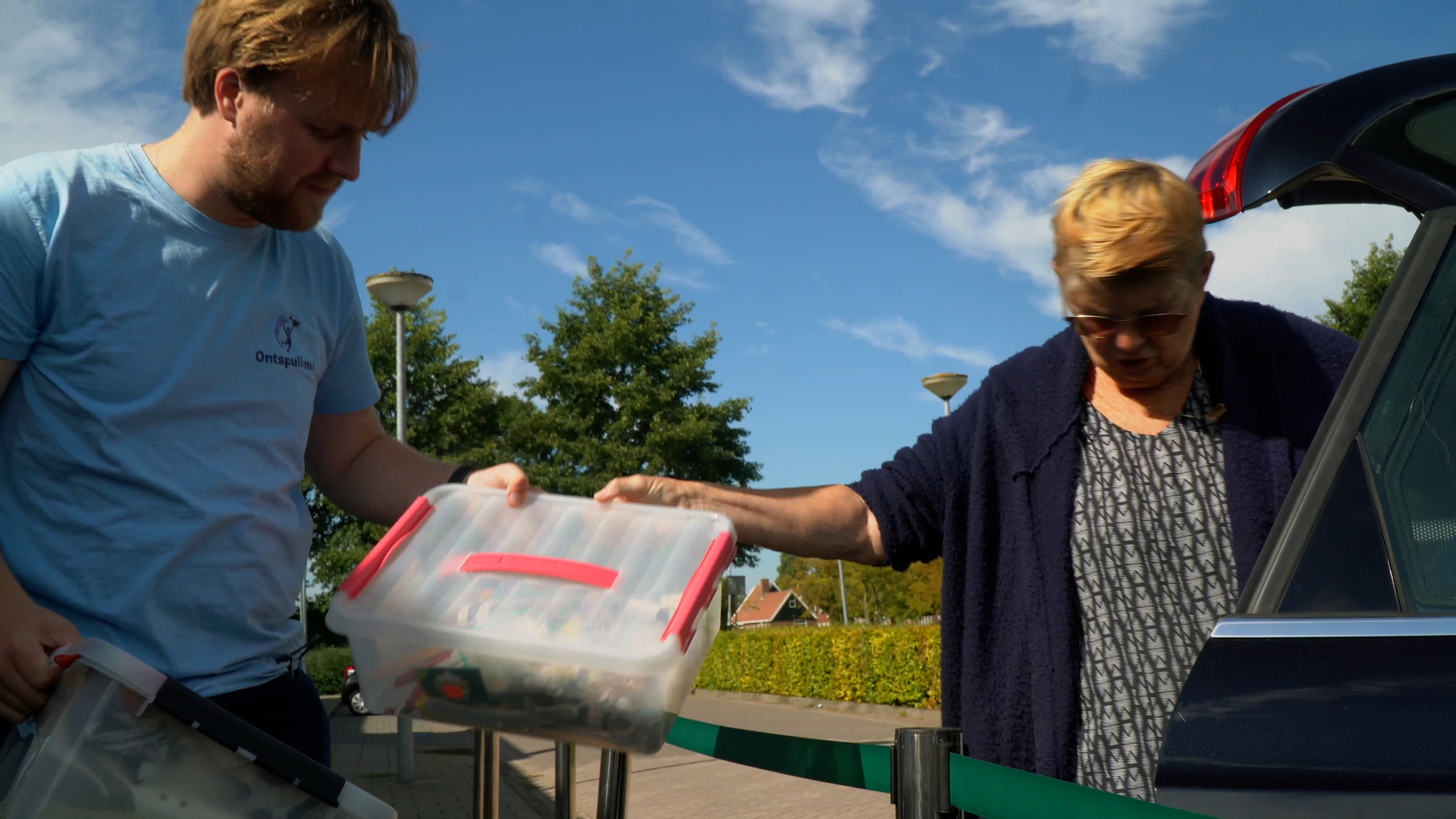 Twee mensen staan buiten, terwijl één van hen een plastic opbergdoos met rode handgrepen vasthoudt. Op de achtergrond zijn bomen en een blauwe lucht zichtbaar.
