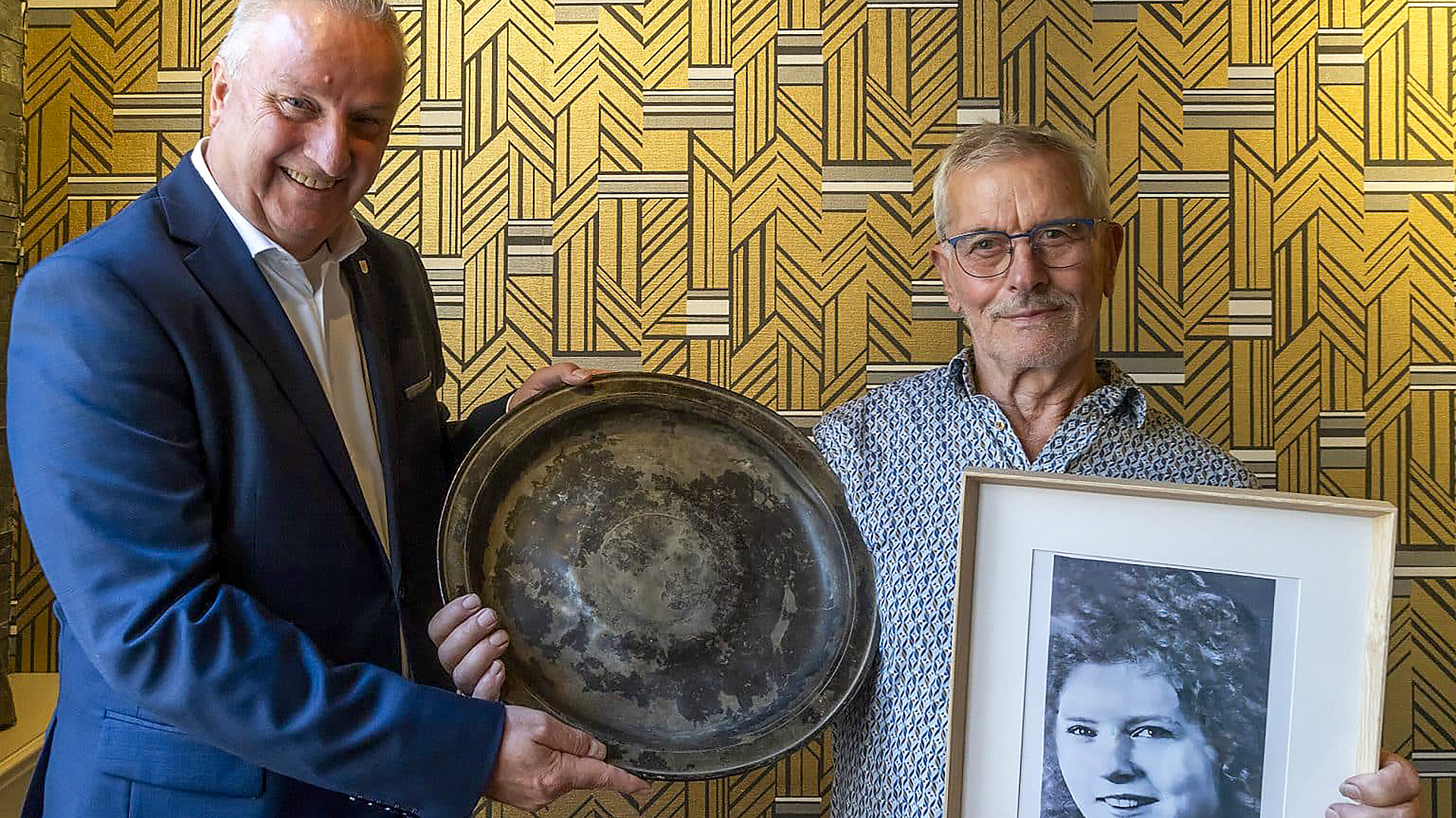 Twee mannen poseren voor een muur met geometrisch patroon; de linker man houdt een grote metalen schaal vast, terwijl de rechter man een ingelijste zwart-witfoto van een vrouw toont.