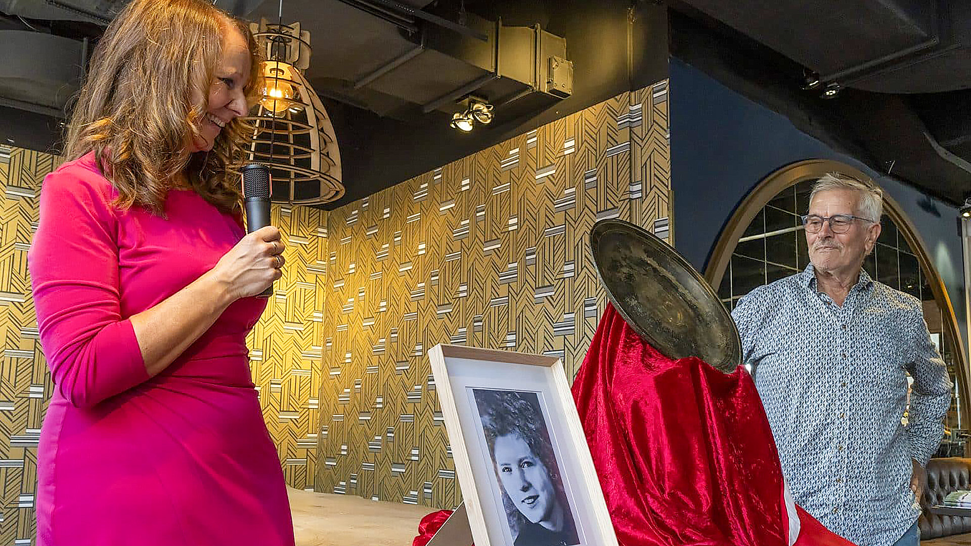 Een vrouw in een roze jurk spreekt in een microfoon terwijl ze naar een ingelijste zwart-witfoto kijkt, naast een man in een blauw shirt en een frisdrankblikje op een rood fluwelen doek in een kamer met geometrisch behang.