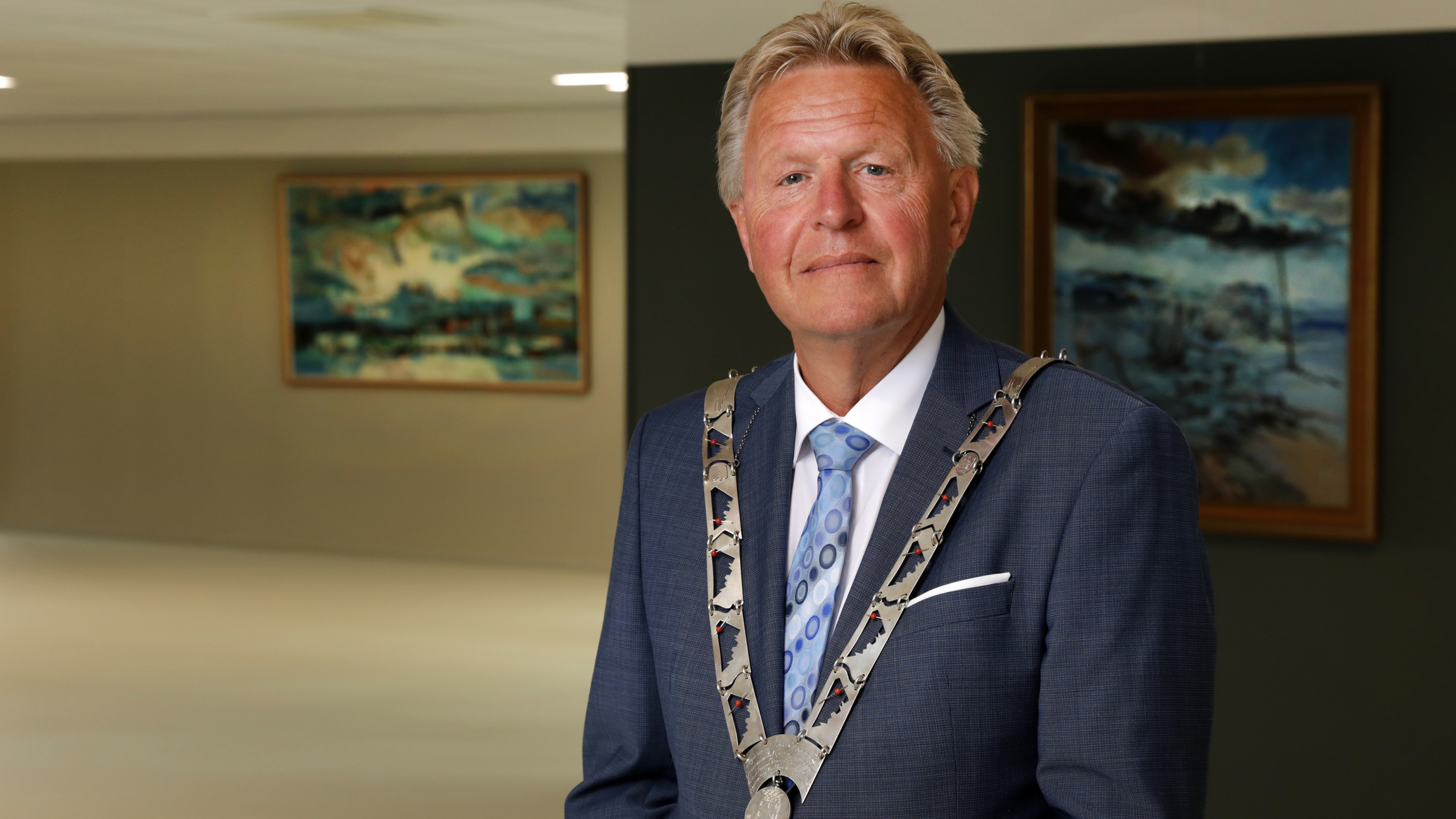 Man in a blue suit wearing a ceremonial chain stands in a room with abstract paintings on the walls.