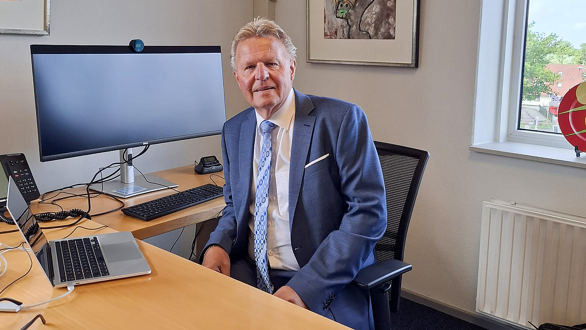 Man in een blauw pak zittend aan een bureau met een laptop en een grote monitor, in een kantoor met een raam en ingelijste kunst aan de muur.