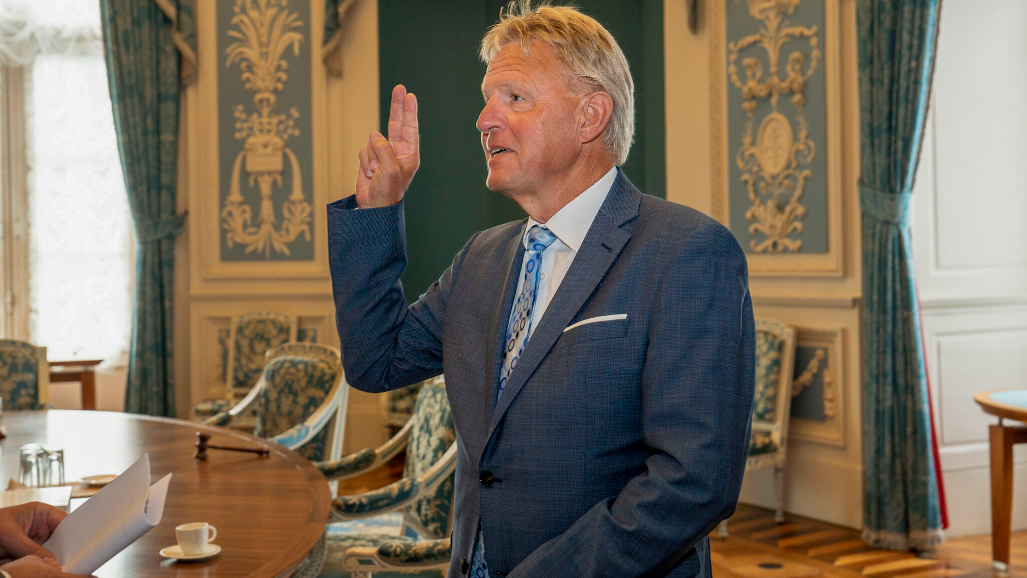 Man in a blue suit taking an oath in an ornate room with blue curtains and patterned walls.