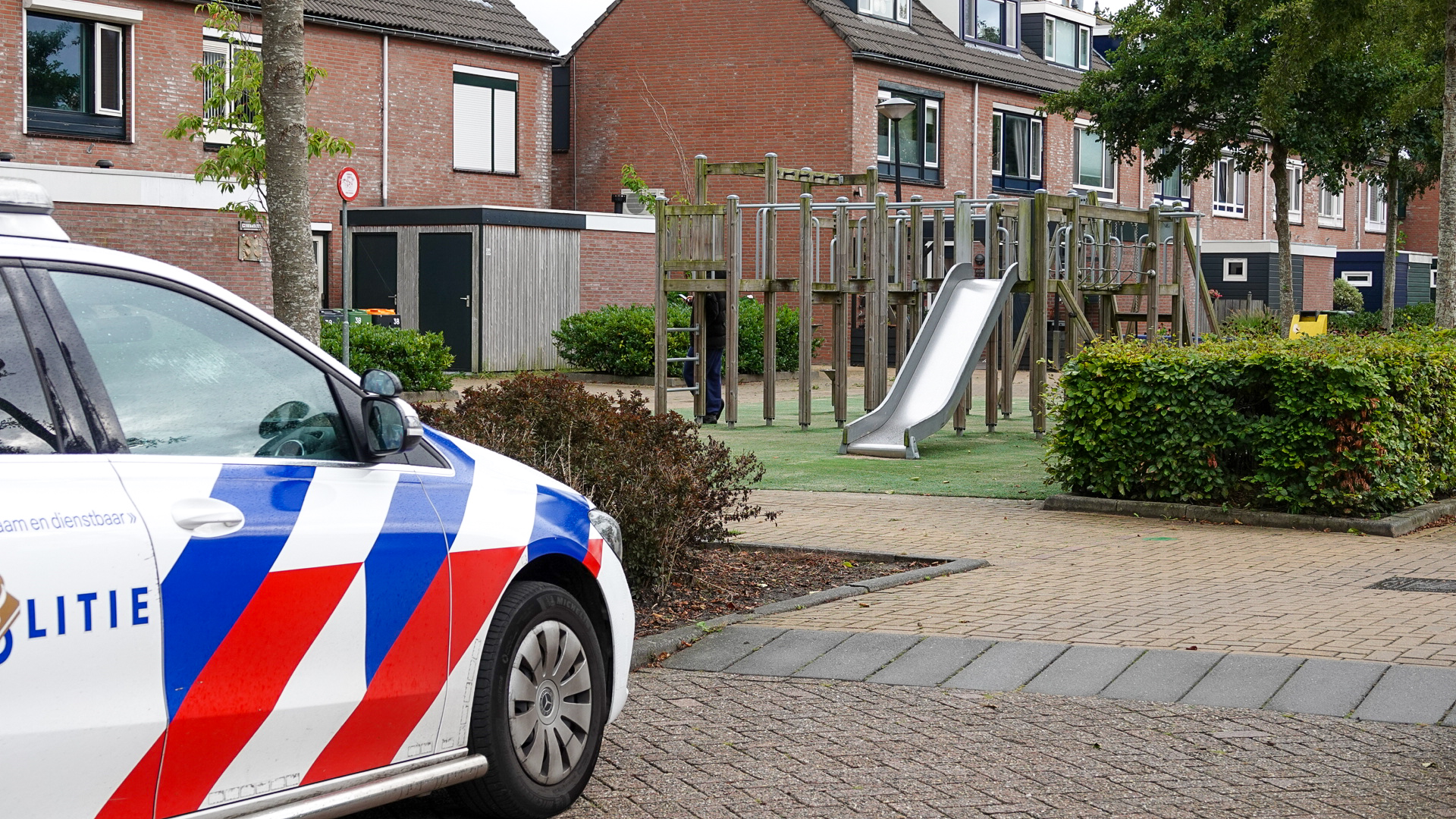 Een politieauto staat geparkeerd naast een speeltuin met glijbaan in een woonwijk met bakstenen huizen.