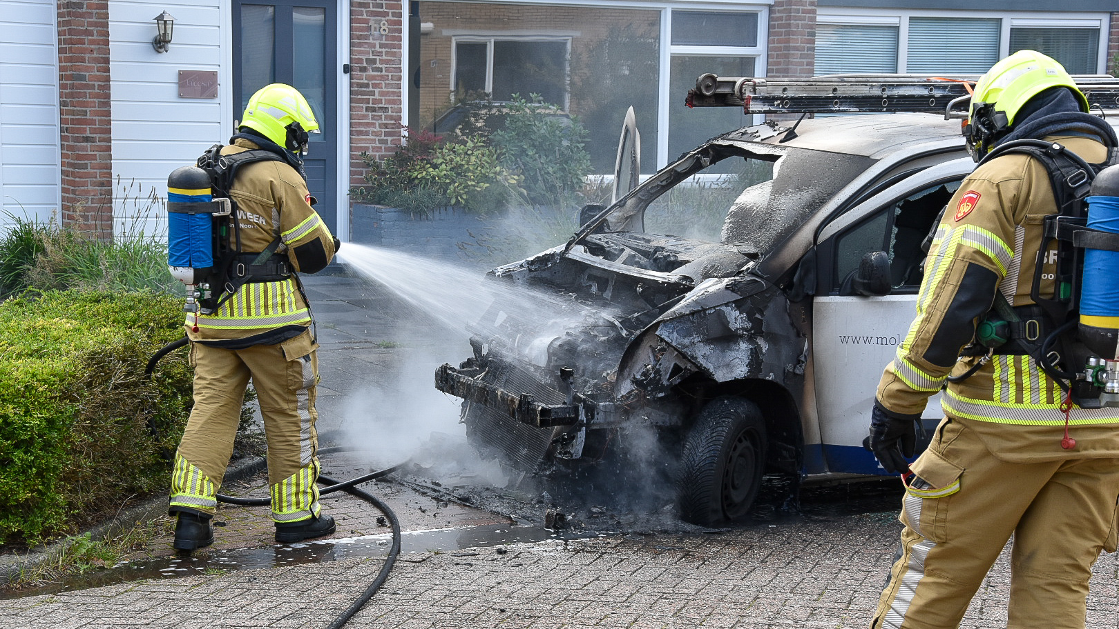 Twee brandweerlieden blussen een uitgebrande bestelwagen op een oprit voor een huis met bakstenen gevel.