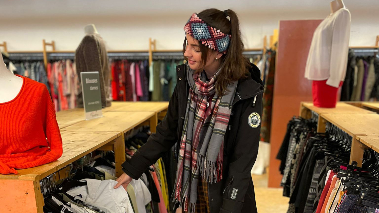 Vrouw met gebreide haarband en sjaal kijkt naar kleding in een winkel, met rekken vol blouses en topjes om haar heen.