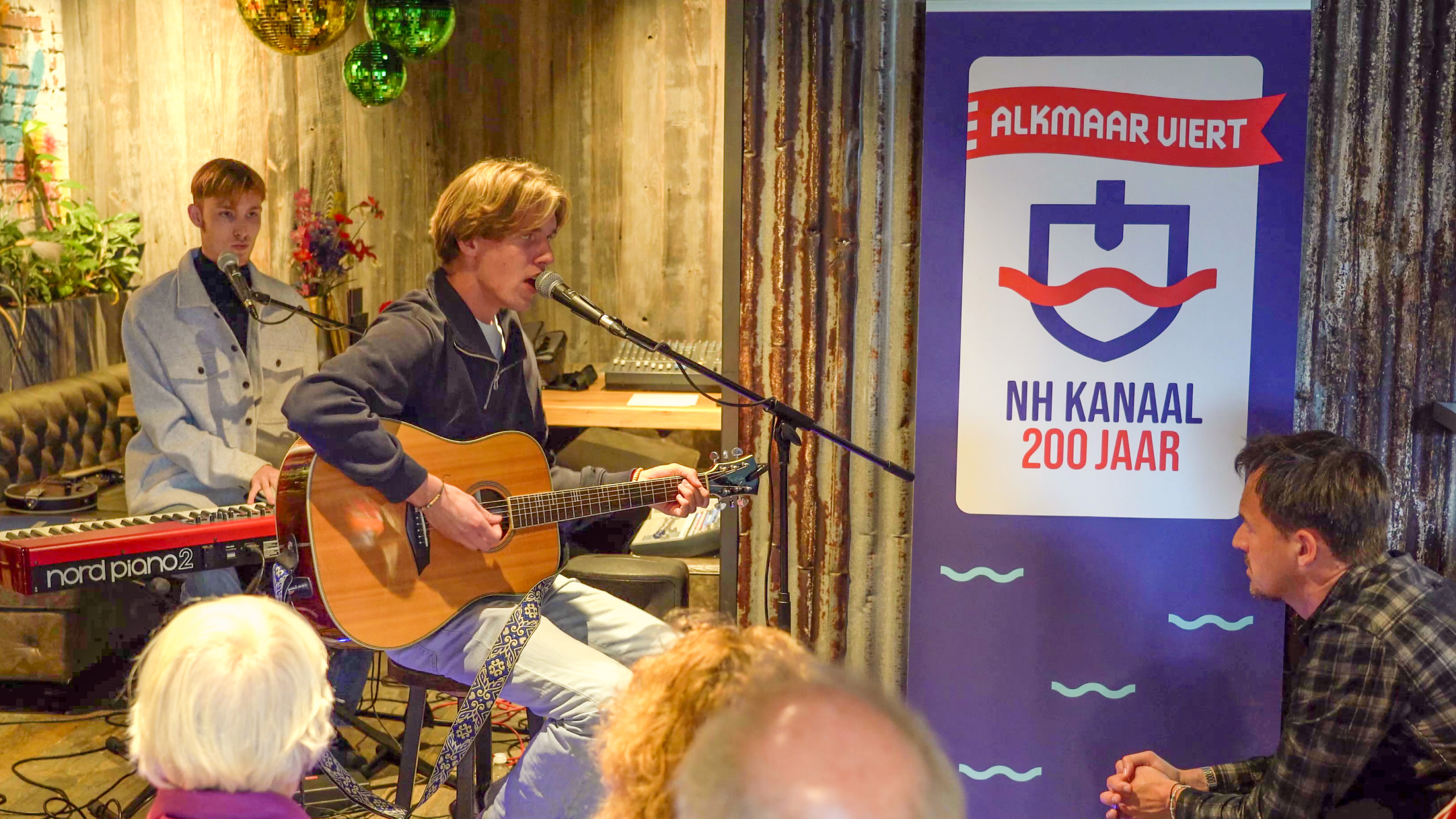 Twee muzikanten treden op tijdens een evenement ter gelegenheid van 200 jaar Noordhollandsch Kanaal, een met een gitaar en de ander achter een keyboard. Een banner met de tekst "Alkmaar viert NH Kanaal 200 jaar" staat naast hen.