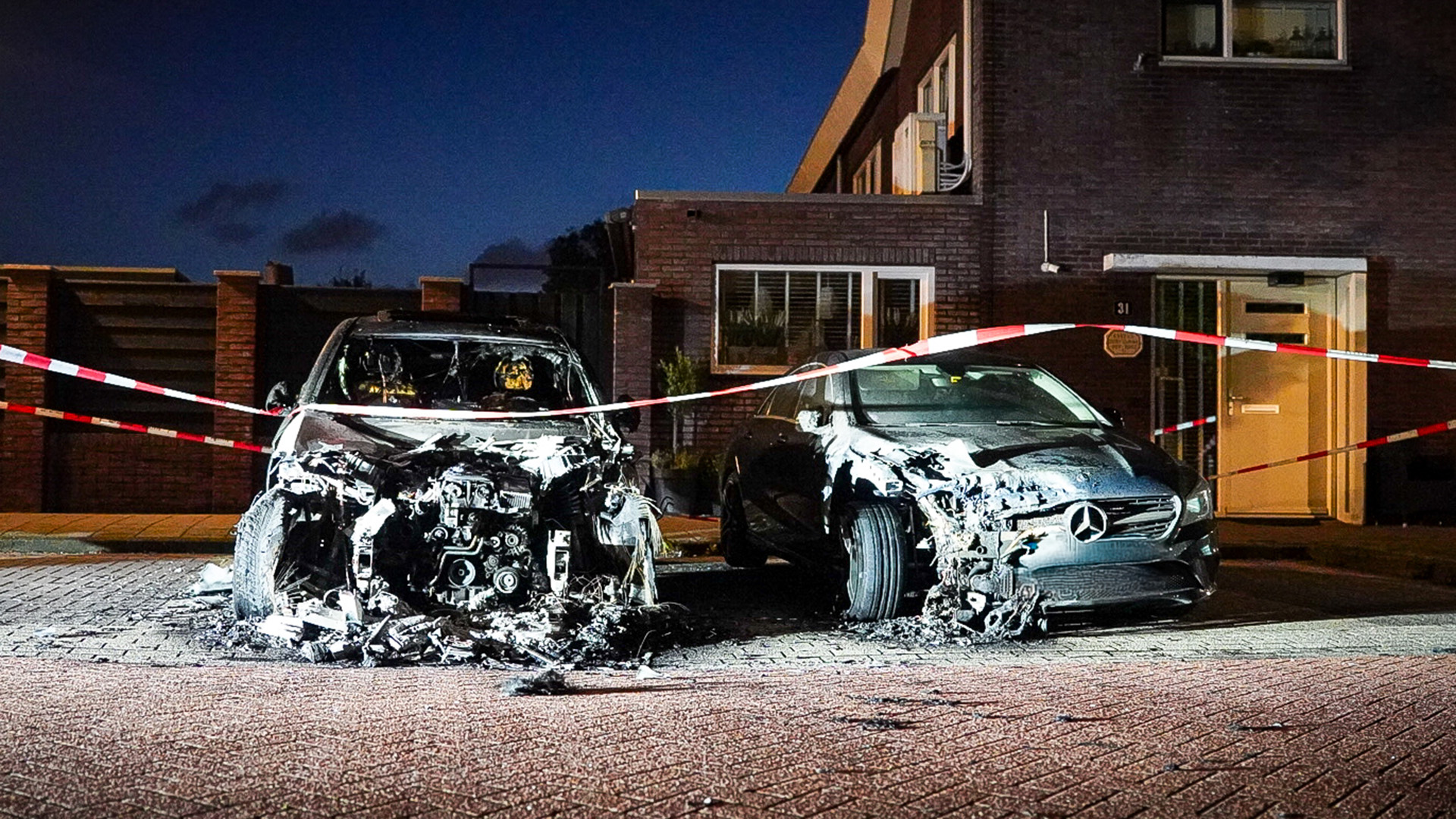 Twee uitgebrande auto's in een woonstraat, omheind met rood-wit afzetlinten, naast een bakstenen gebouw.