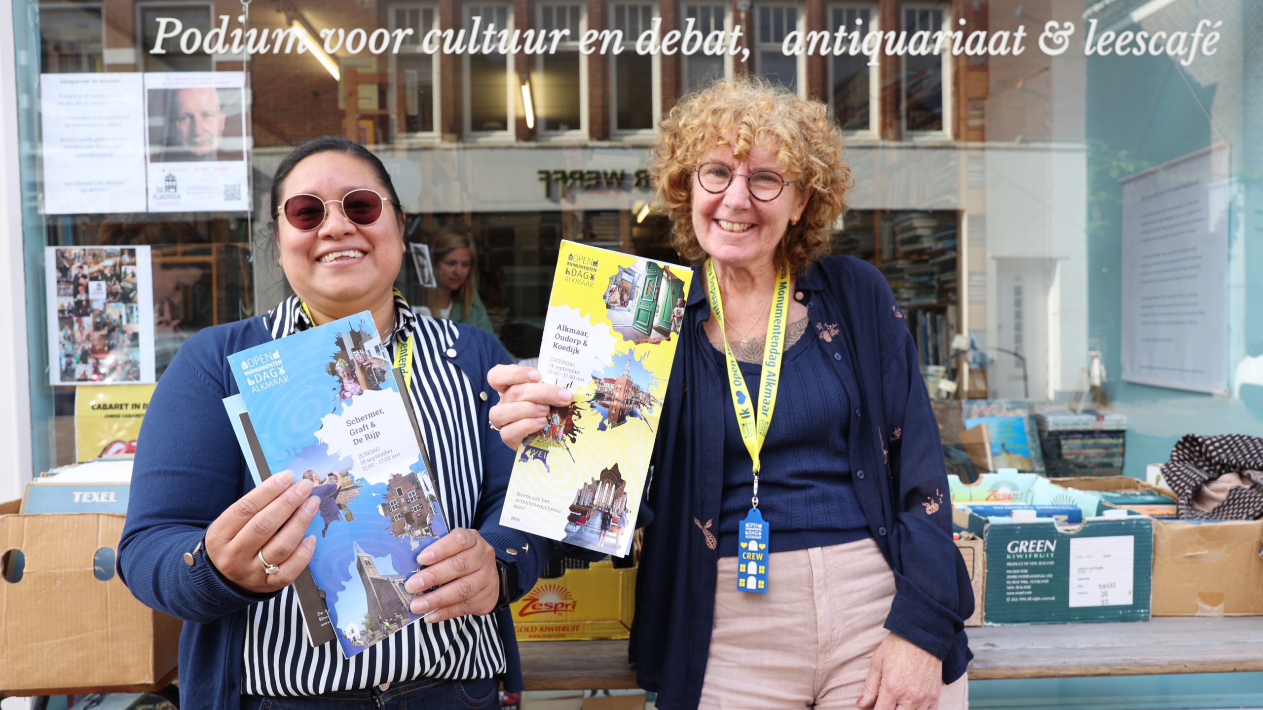 Twee lachende personen staan voor een gevel met de tekst 'Podium voor cultuur en debat, antiquariaat & leescafé', en tonen folders van Open Monumentendag Alkmaar.